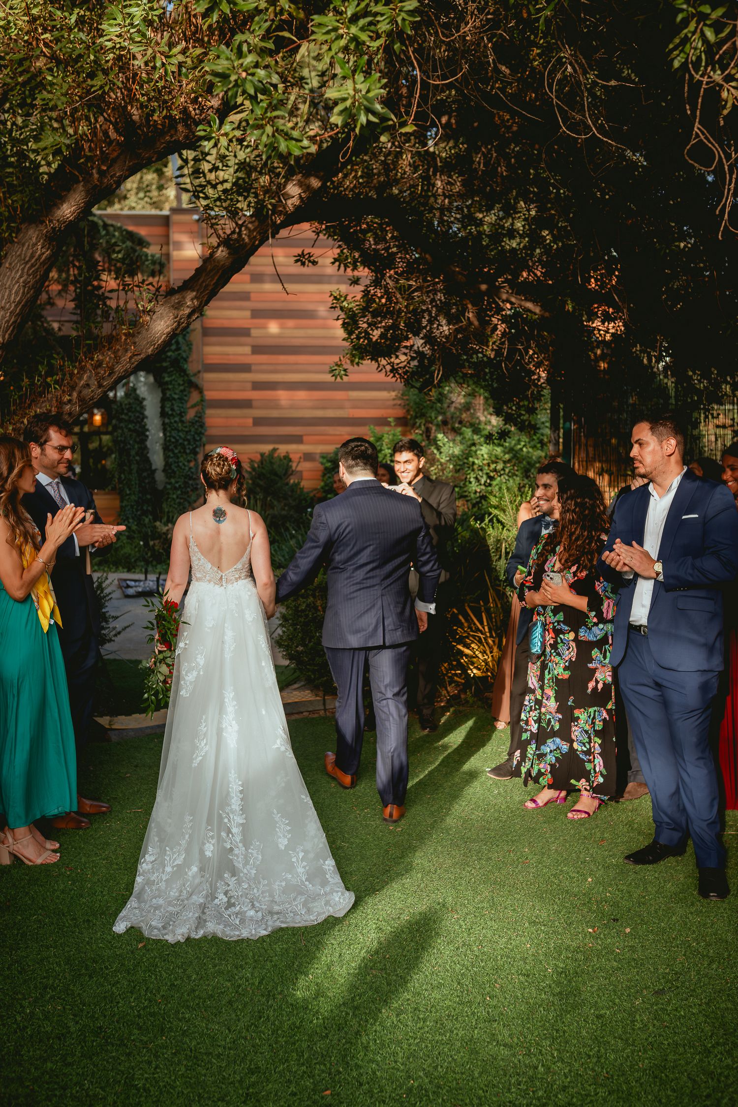 Bride and groom walk through cheering guests under trees at elegant nighttime outdoor wedding reception.