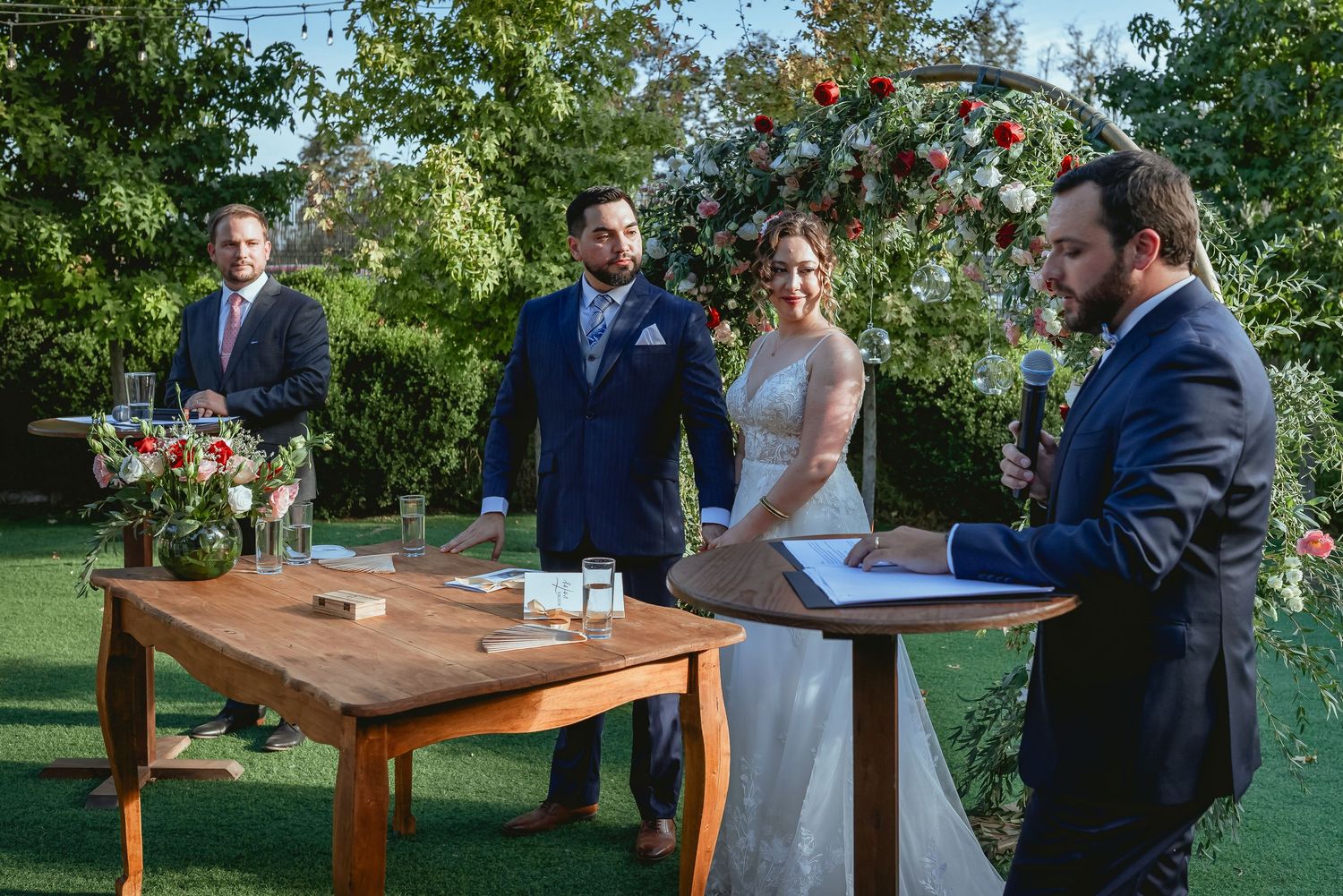 Officiant reads ceremony script as bride and groom stand before floral arch with witnesses.