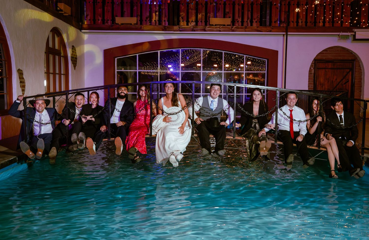 Bride, groom, and wedding guests sit together with feet in pool at night reception.