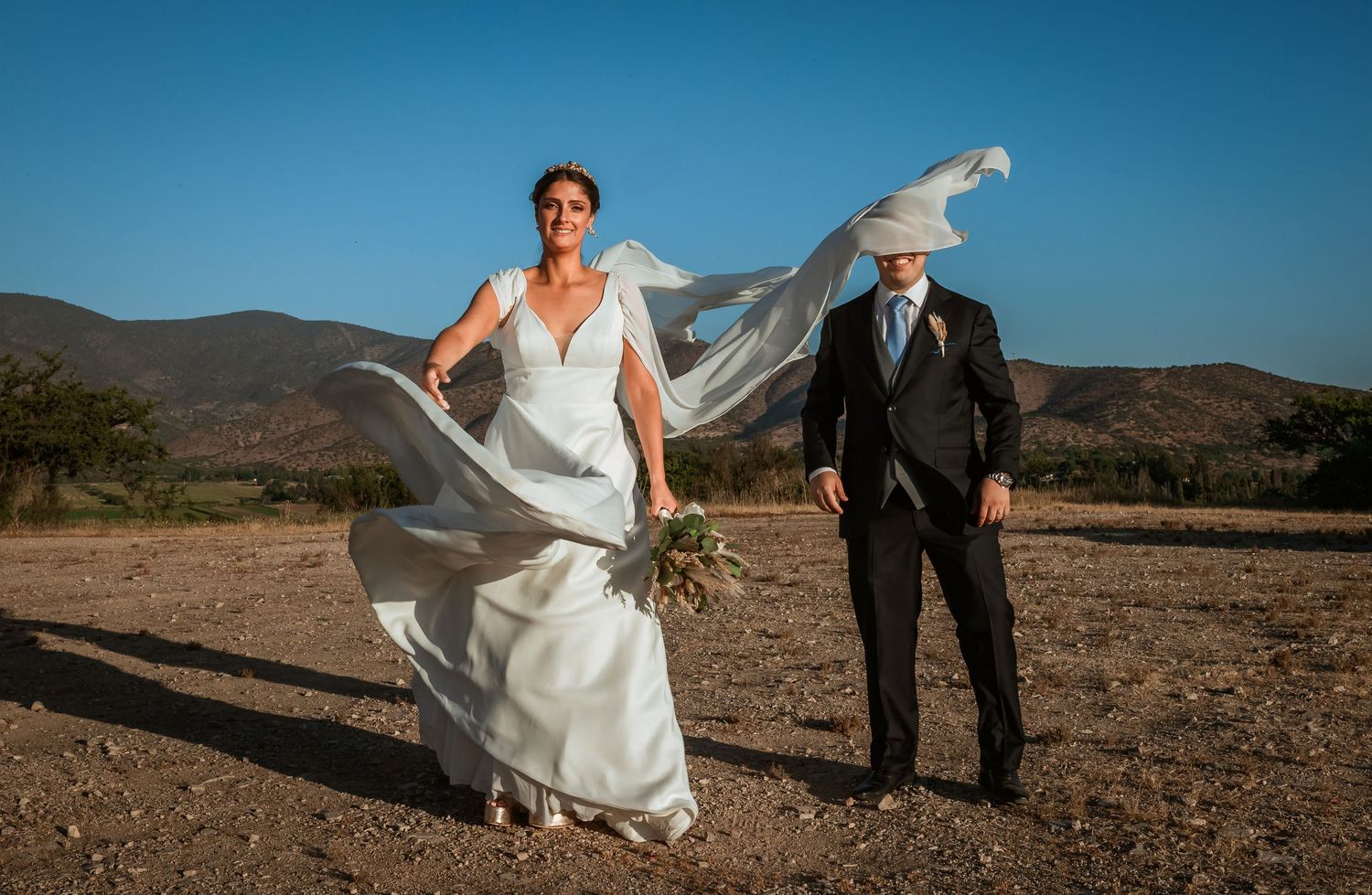 Elegant bride and groom stand in arid landscape as her cathedral veil billows in the breeze.