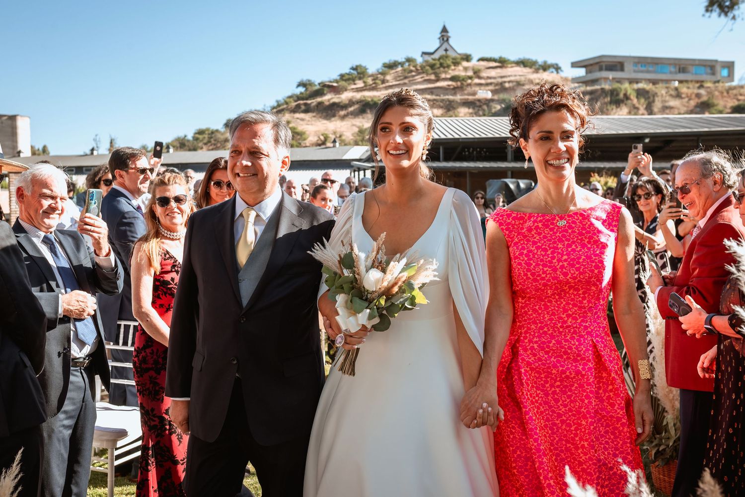 Radiant bride walks with parents down sunny outdoor wedding ceremony aisle.