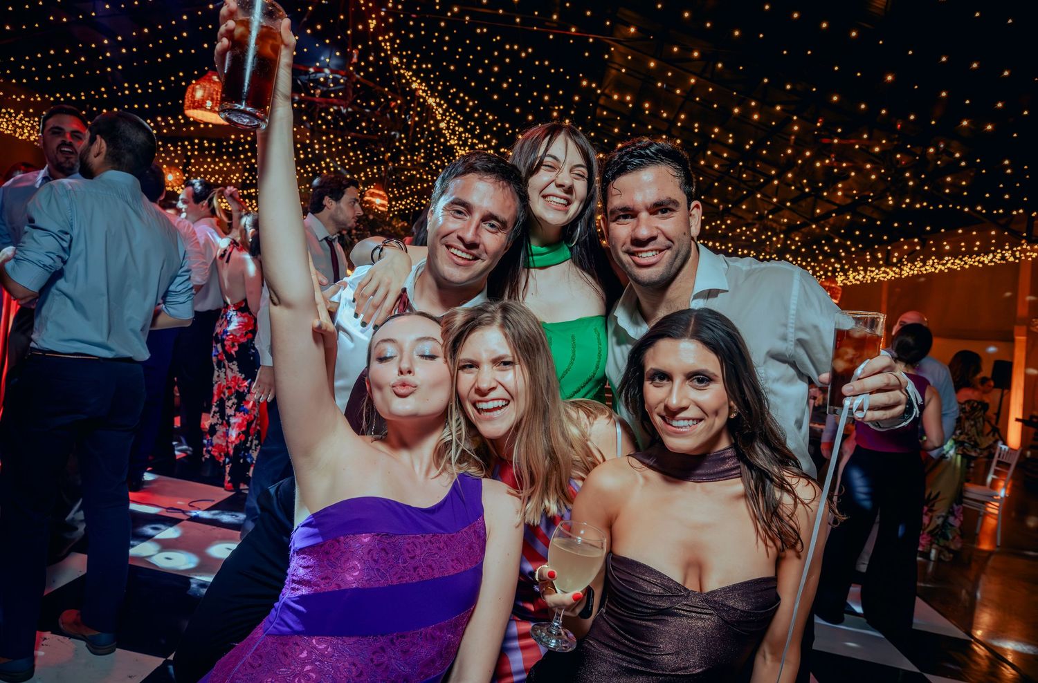Happy group of wedding guests smile together under warm string lights at reception.