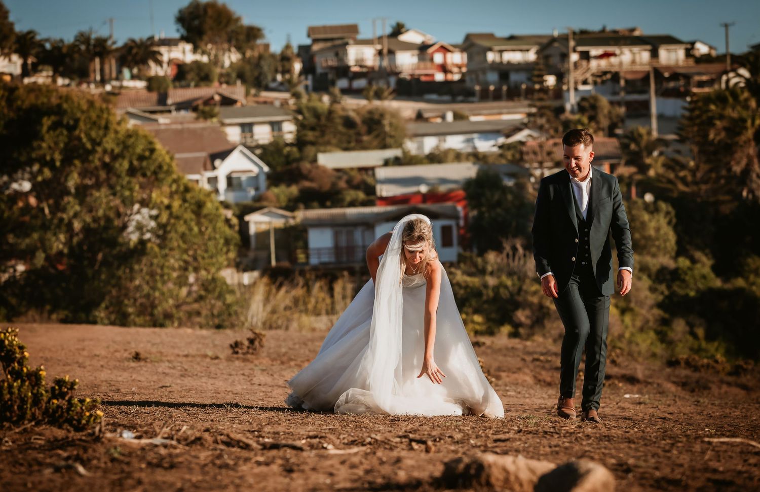 Bride and groom walking on a dusty hillside path with a residential neighborhood behind.