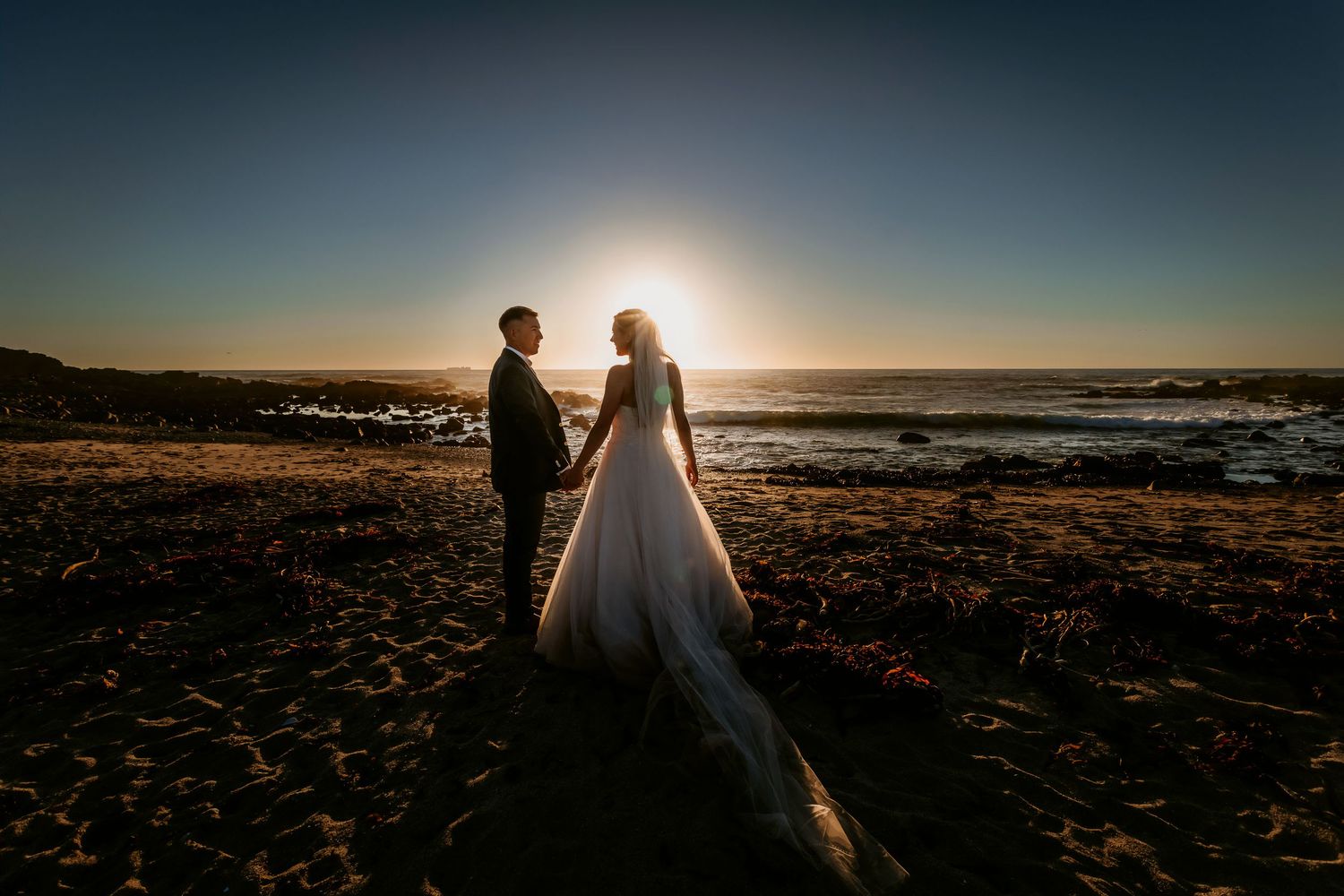 Bride and groom facing each other on beach, backlit by stunning golden sunset over ocean.