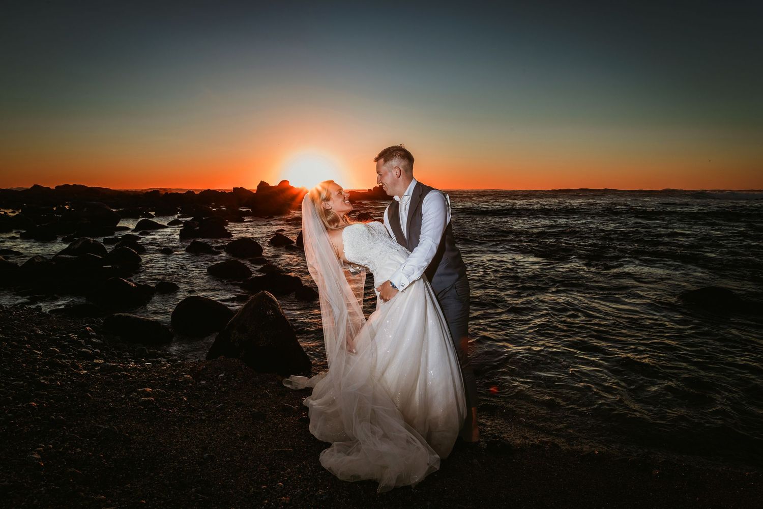 Groom dipping bride romantically on rocky coast with deep red and orange sunset glow.
