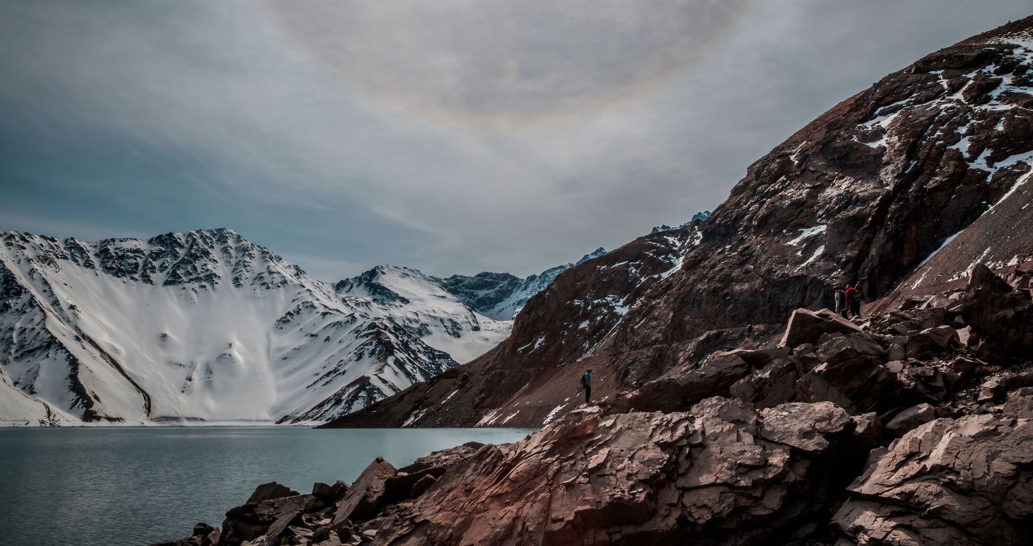 Rocky foreground leads to serene glacial lake surrounded by snow-covered mountains under dramatic sky.