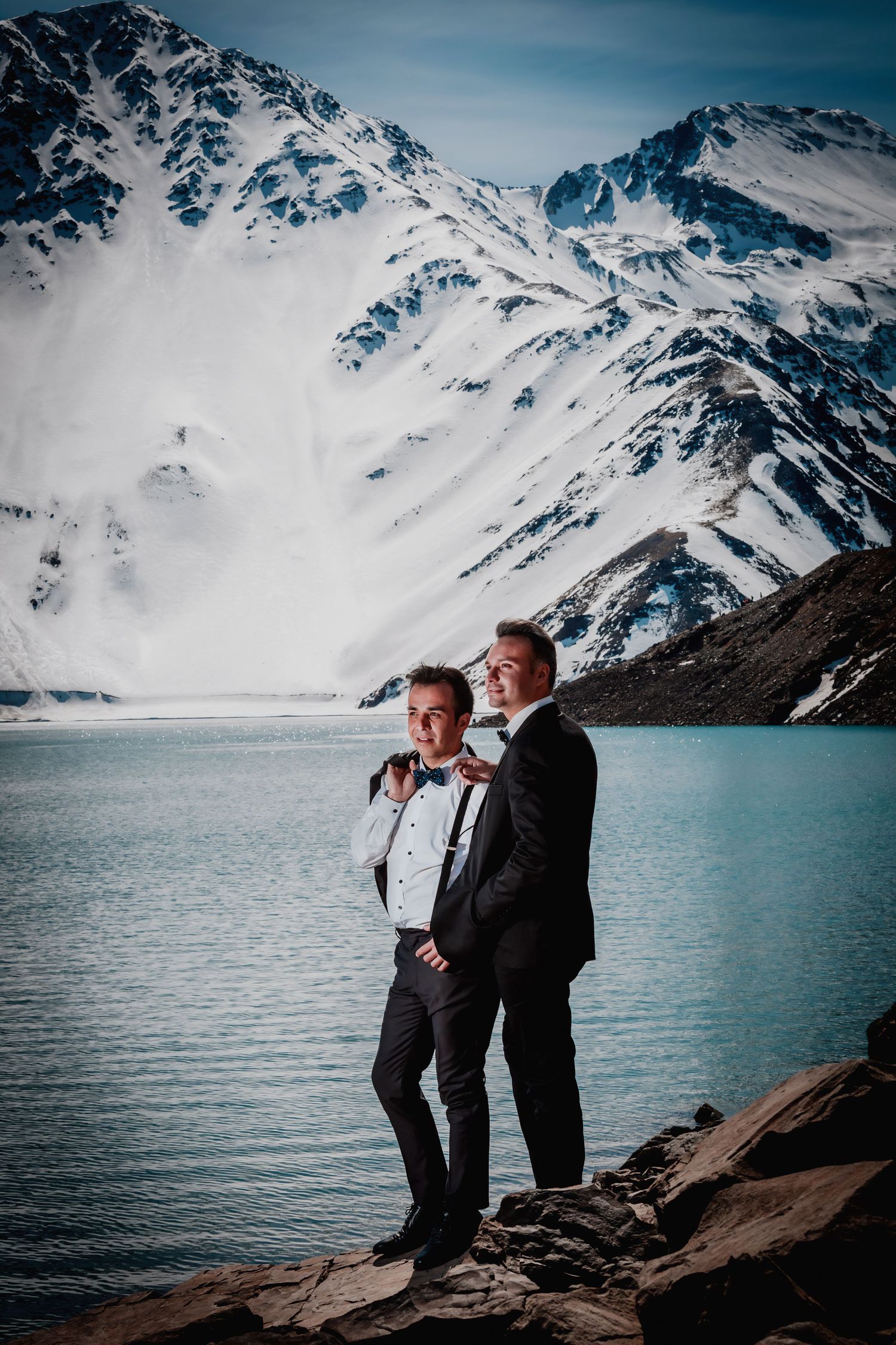 Same-sex couple in tuxedos holding hands by a stunning turquoise alpine lake with snowy peaks.