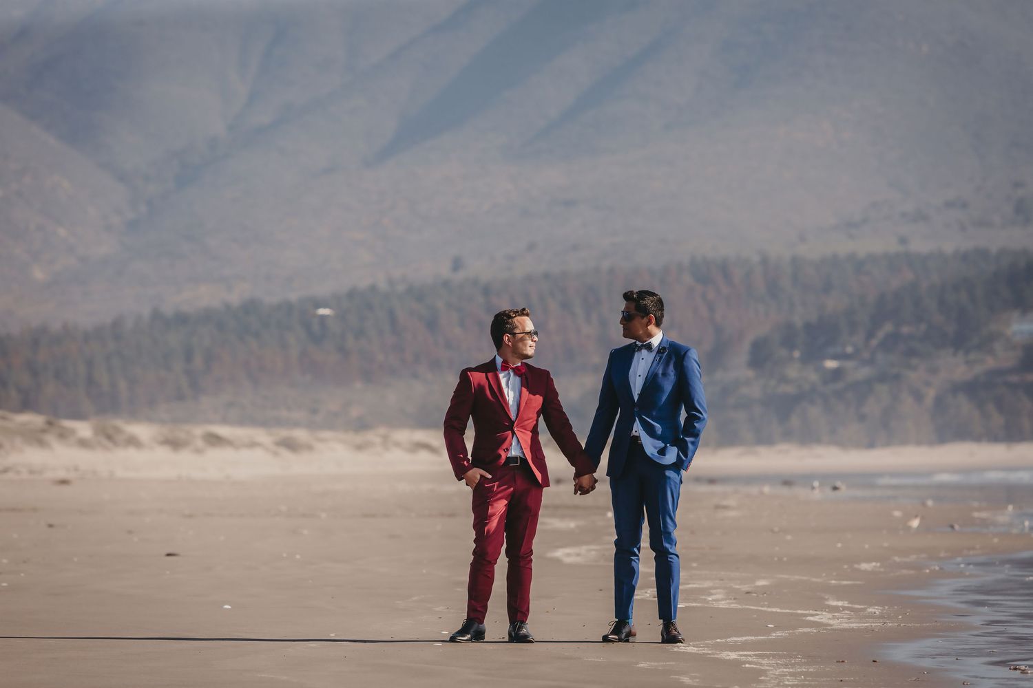 Two grooms holding hands and walking along a scenic beach with mountains in the background.