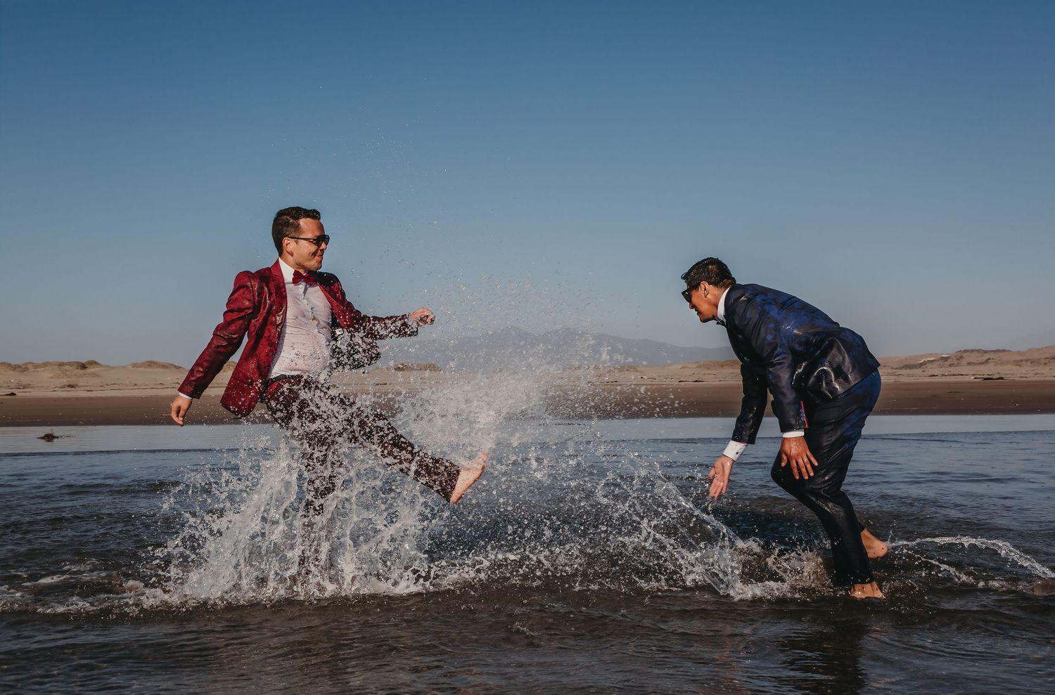 Two grooms splash water at each other on a beach, one in a red suit and one in a dark wetsuit.