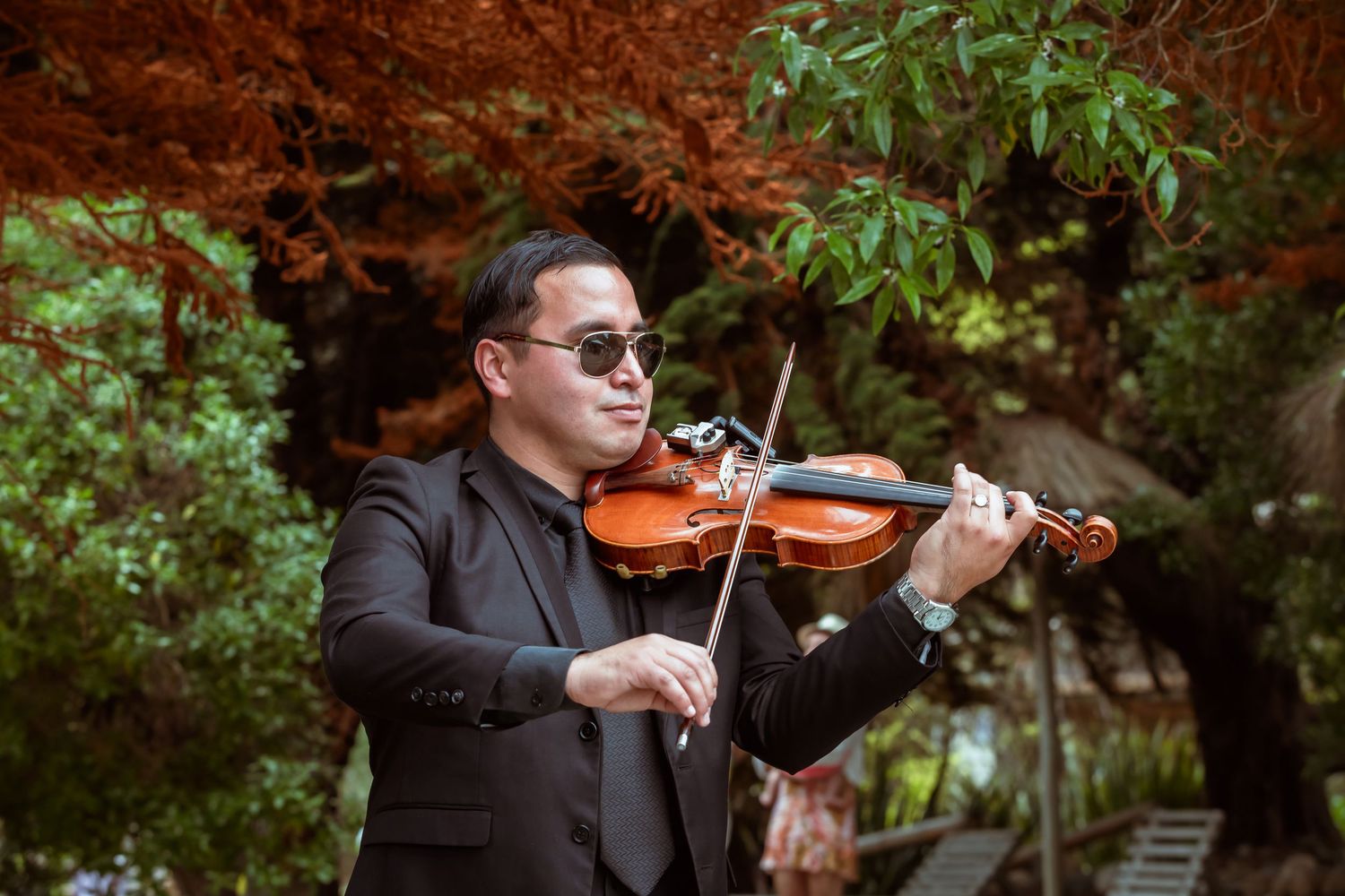 A violinist in a black suit and sunglasses plays violin outdoors among autumn foliage.