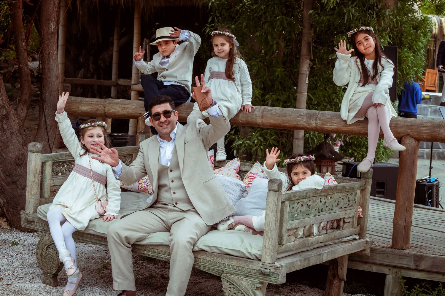 Groom posing playfully with children in white wedding attire on rustic bench.