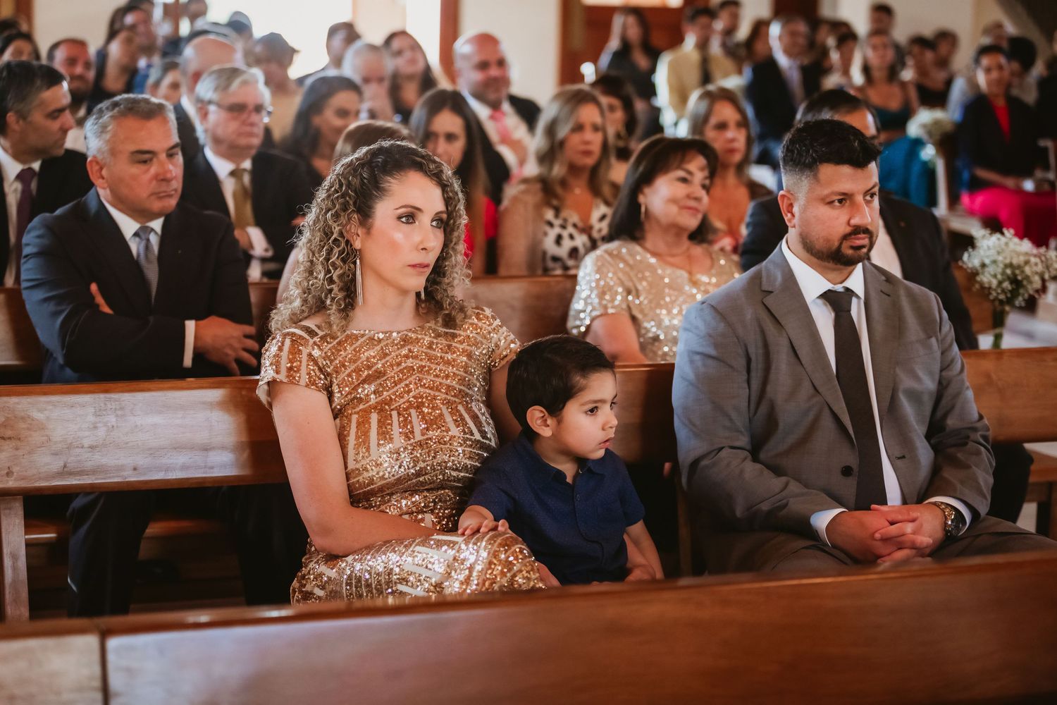 Wedding guests seated in church pews including woman in gold dress with young boy.