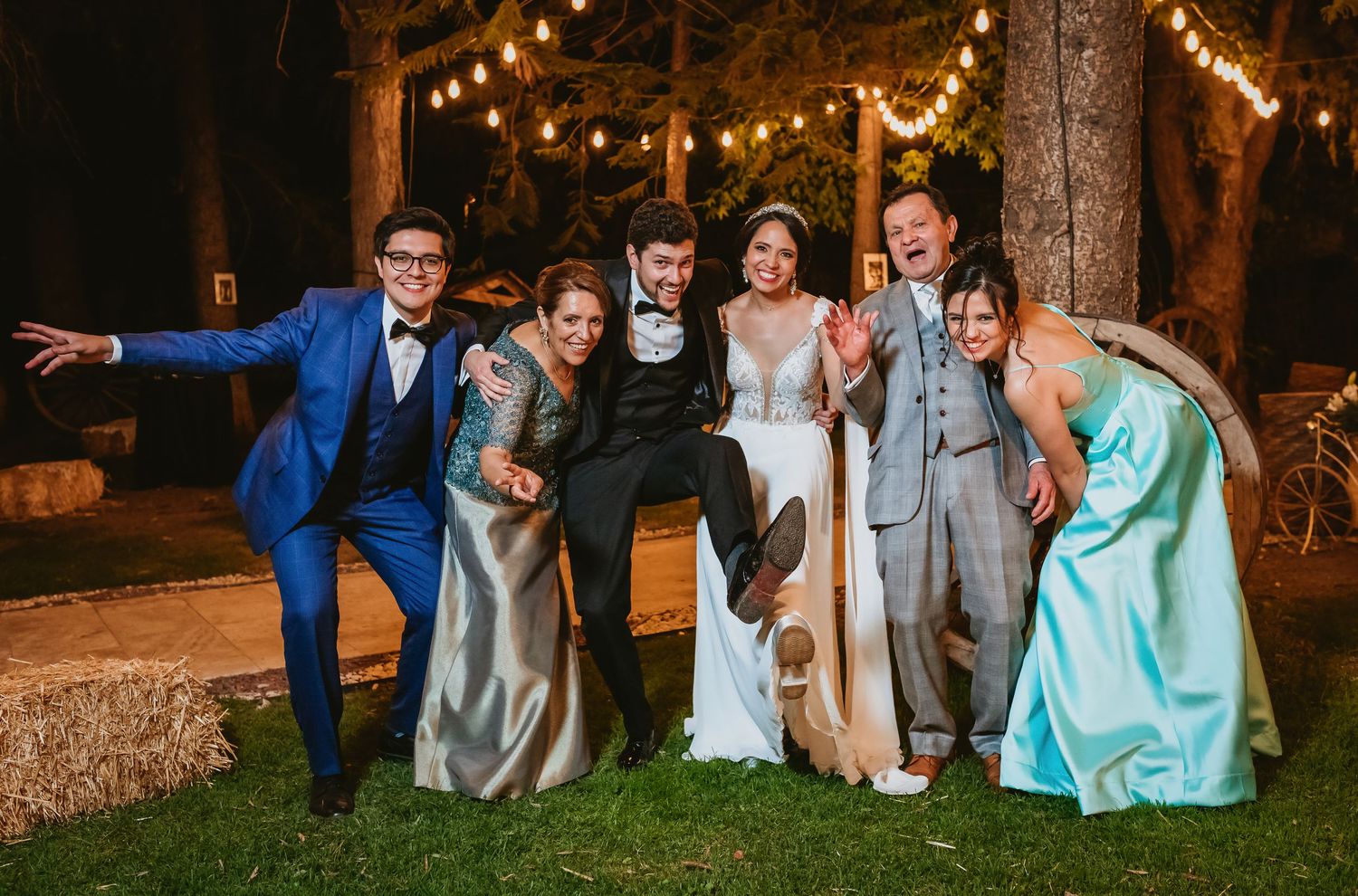 Joyful wedding party group laughing and posing outdoors at night under warm string lights.