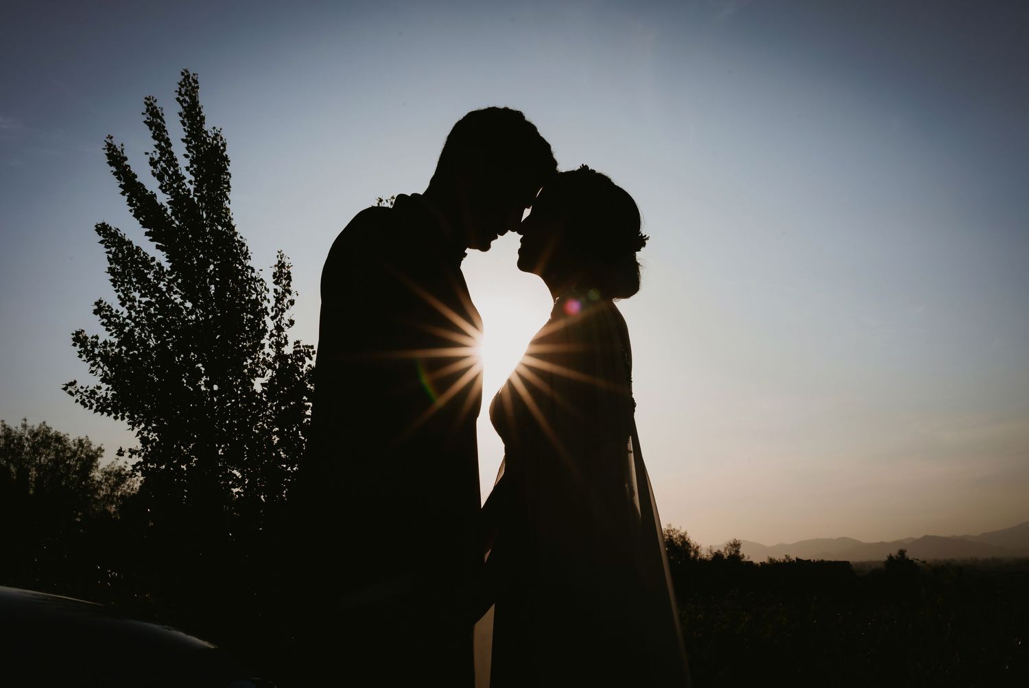 Romantic silhouette of bride and groom touching foreheads at sunset with sunburst.