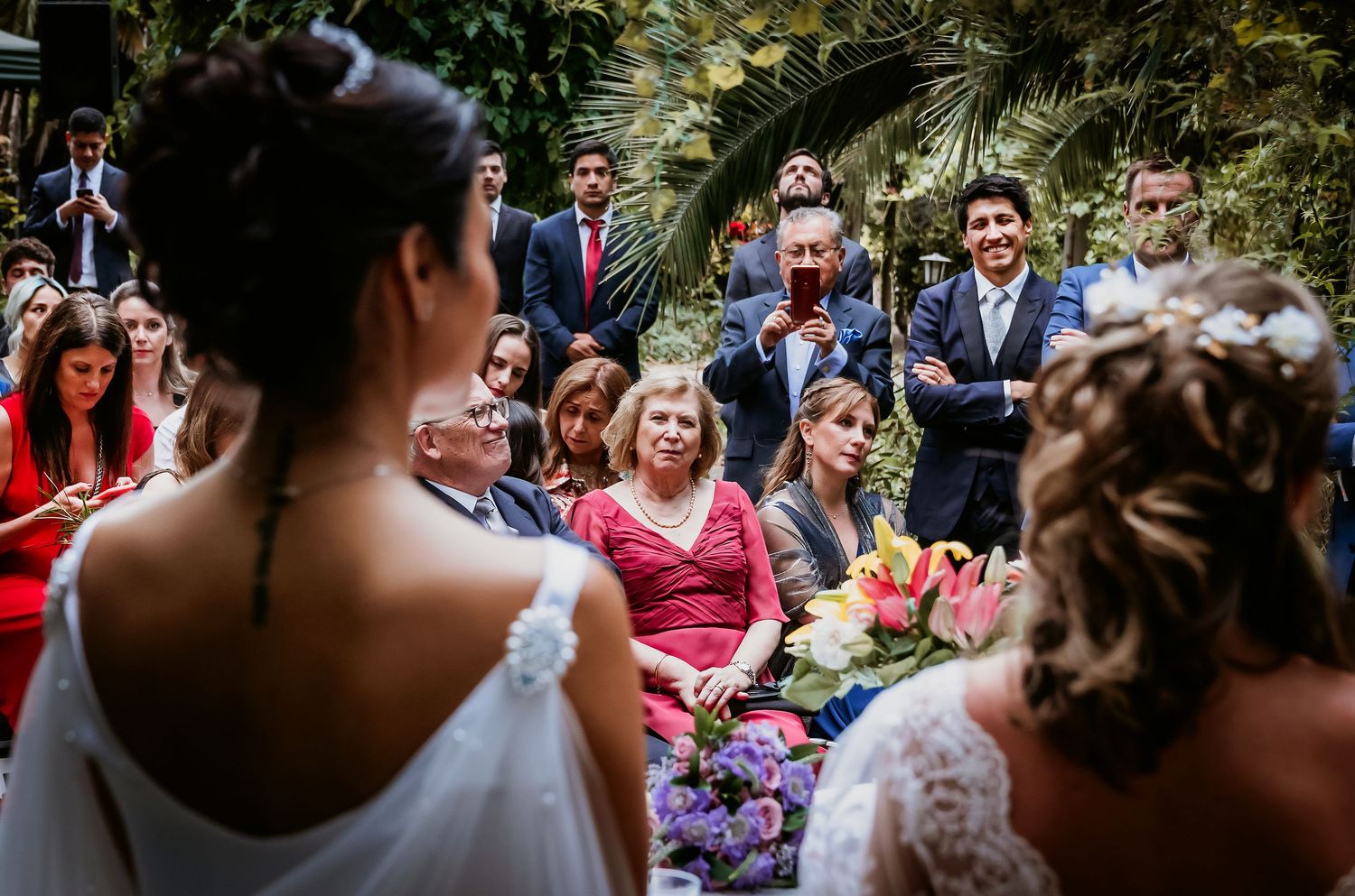 Two brides stand before smiling wedding guests during an outdoor garden ceremony.