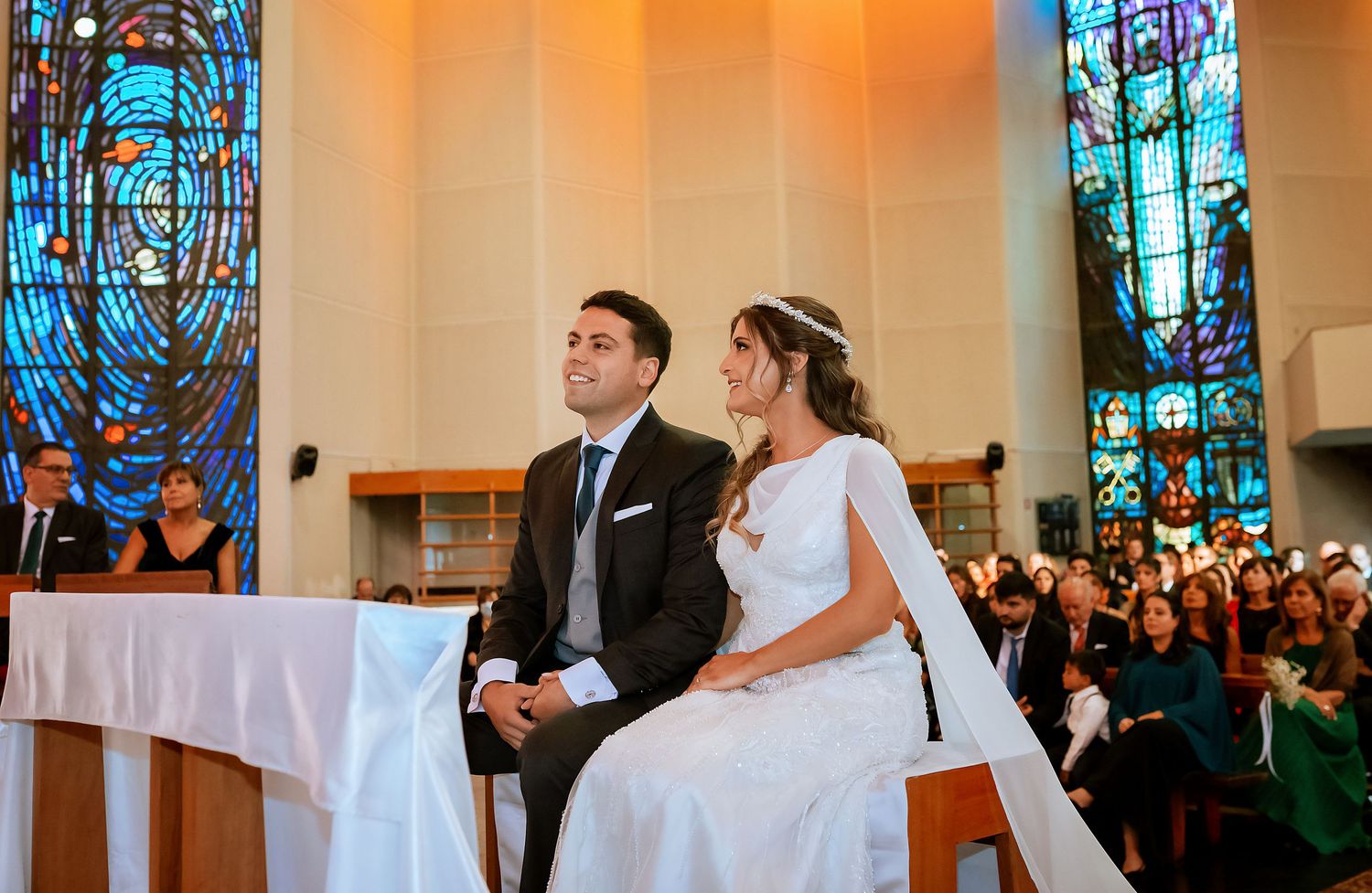 Bride and groom seated together at church altar during wedding ceremony with colorful stained glass windows behind.