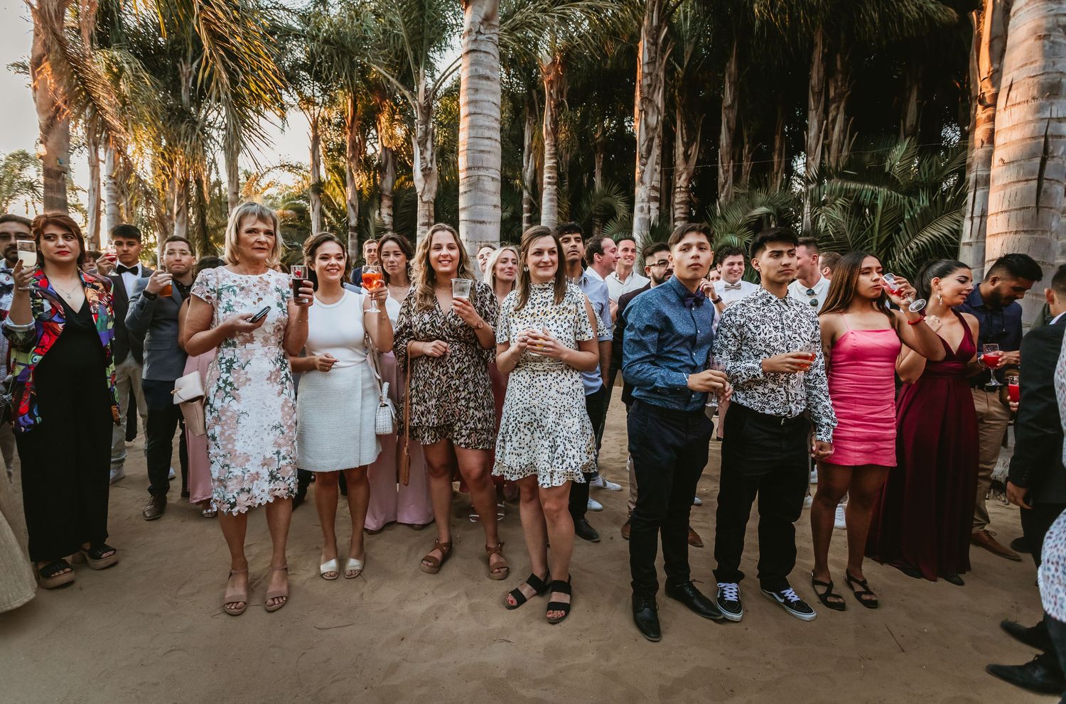 Wedding guests clap and celebrate outdoors under palm trees at a tropical beach wedding reception.