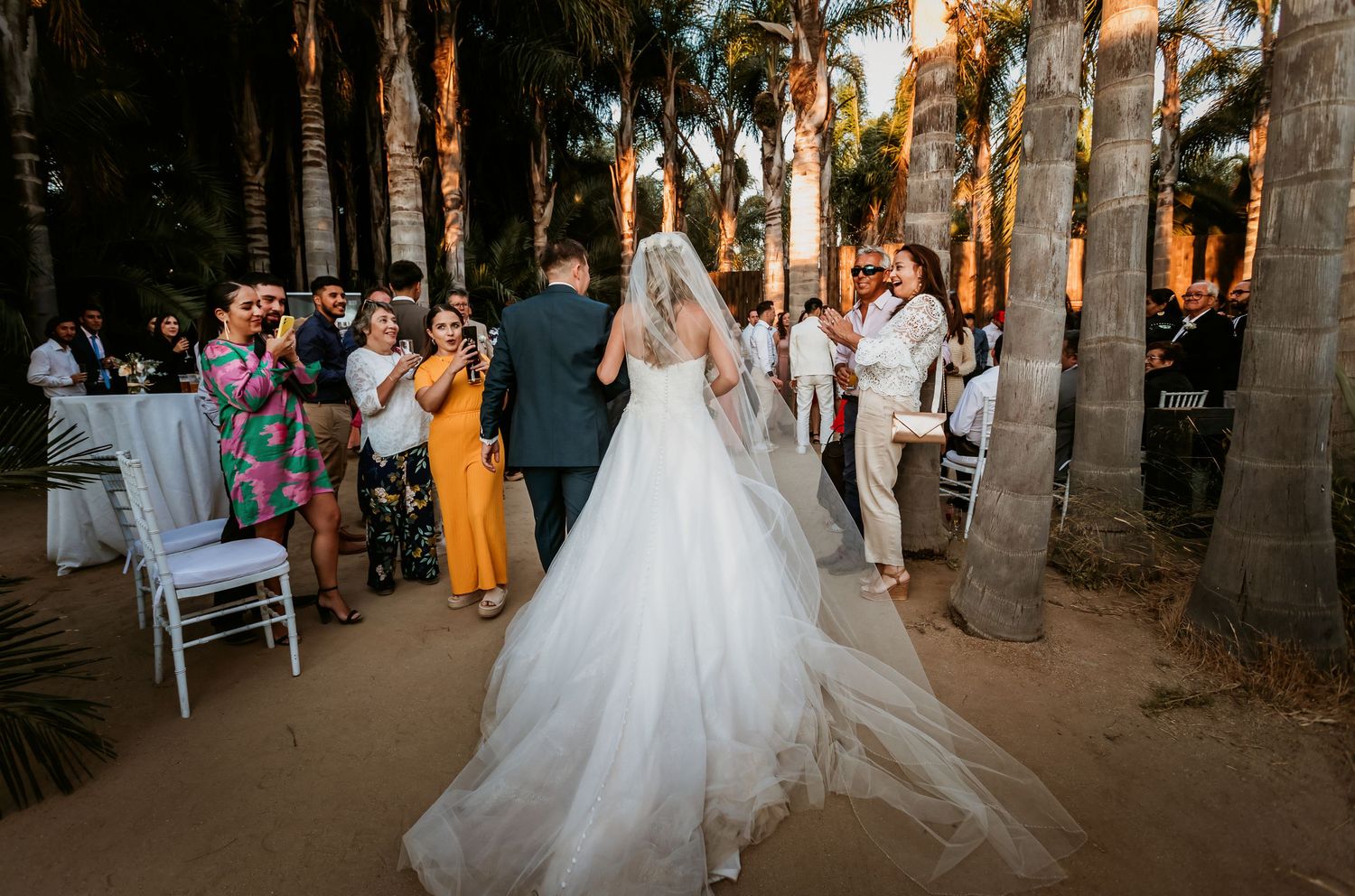 Bride in white ballgown walks with groom through palm tree wedding ceremony while guests celebrate.