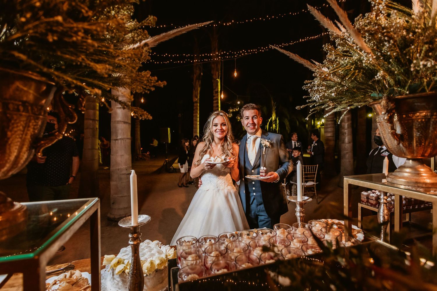 Bride and groom smile together at dessert table with candles at outdoor wedding reception.