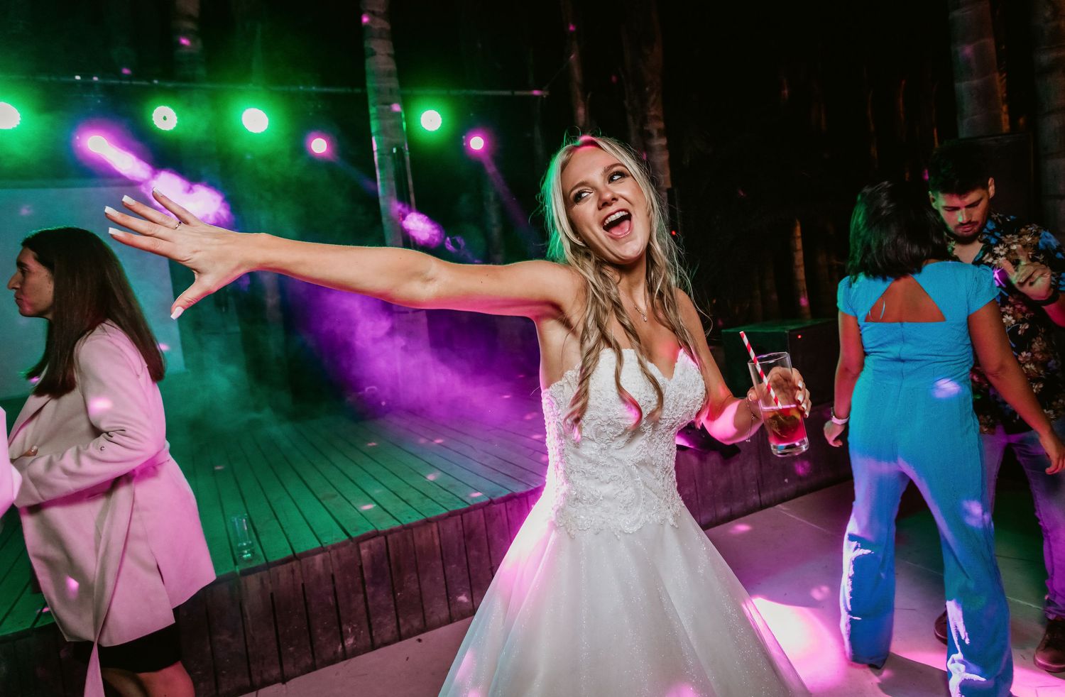 Radiant bride in white gown dances joyfully under colorful lights at lively wedding reception.
