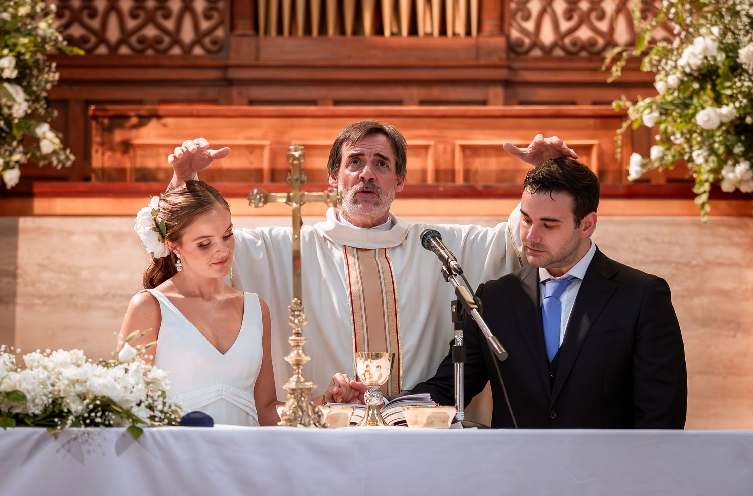 Catholic priest raises hand in blessing over bride and groom at decorated altar during wedding mass.