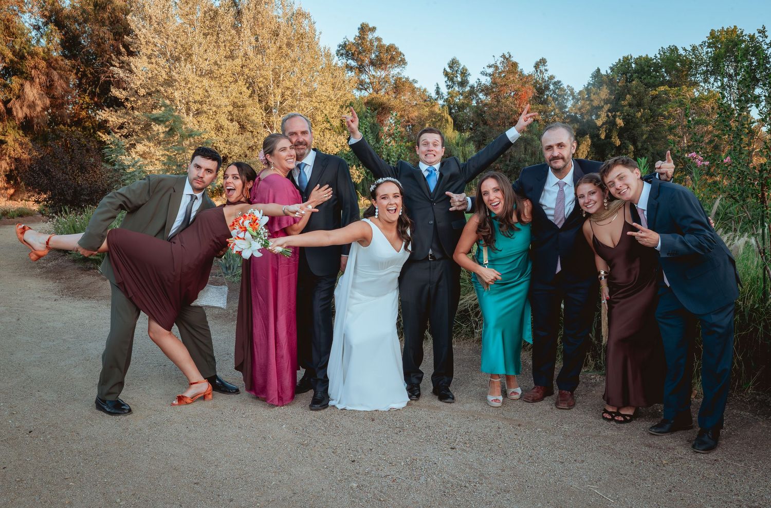 Bride and groom celebrate with wedding party in colorful attire outdoors in autumn setting.