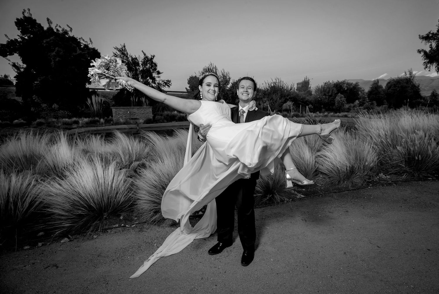 Black and white photo of groom carrying laughing bride with arms outstretched outdoors.