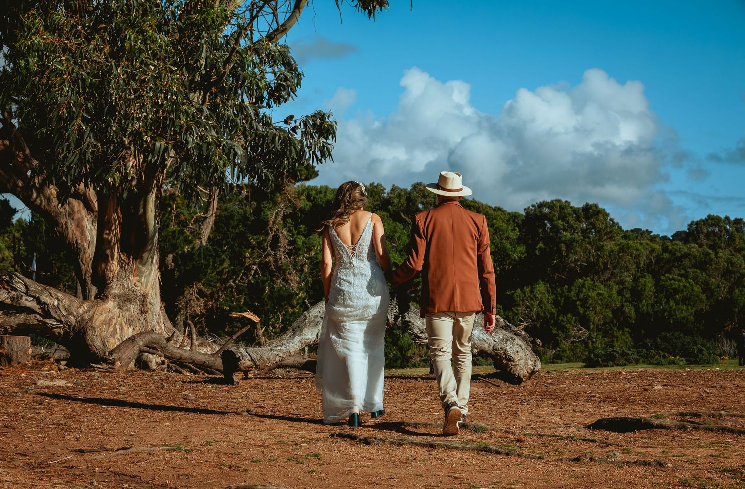 Couple holding hands walk away across red dirt toward large gum trees on a sunny day, she in a white dress, he in a rust shirt and hat.