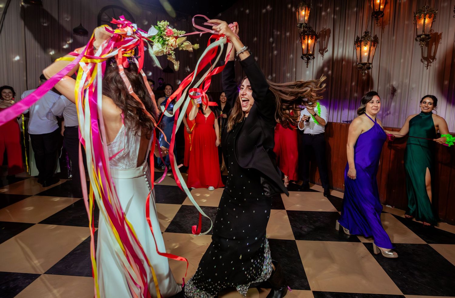 Bride and female guest dance with colorful ribbons on checkered wedding dance floor.