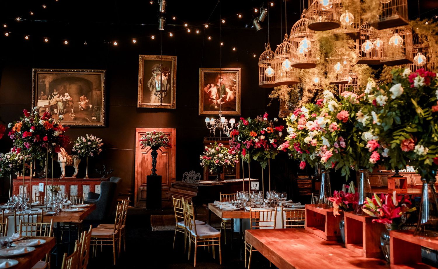 Romantic wedding venue with dark walls, framed paintings, tall floral arrangements, and string lights.