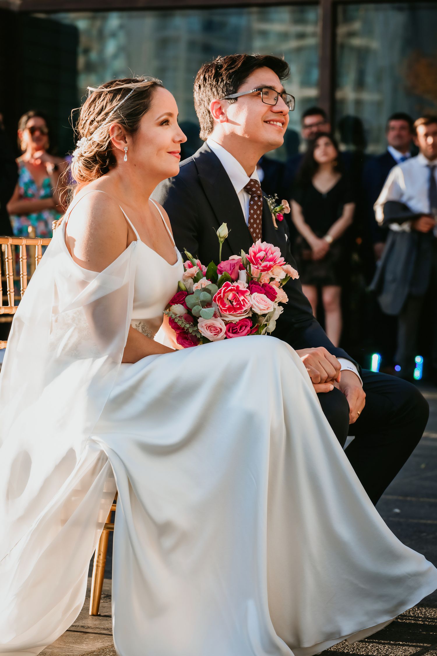 Radiant bride in white gown and groom in dark suit sit together at sunny wedding ceremony.