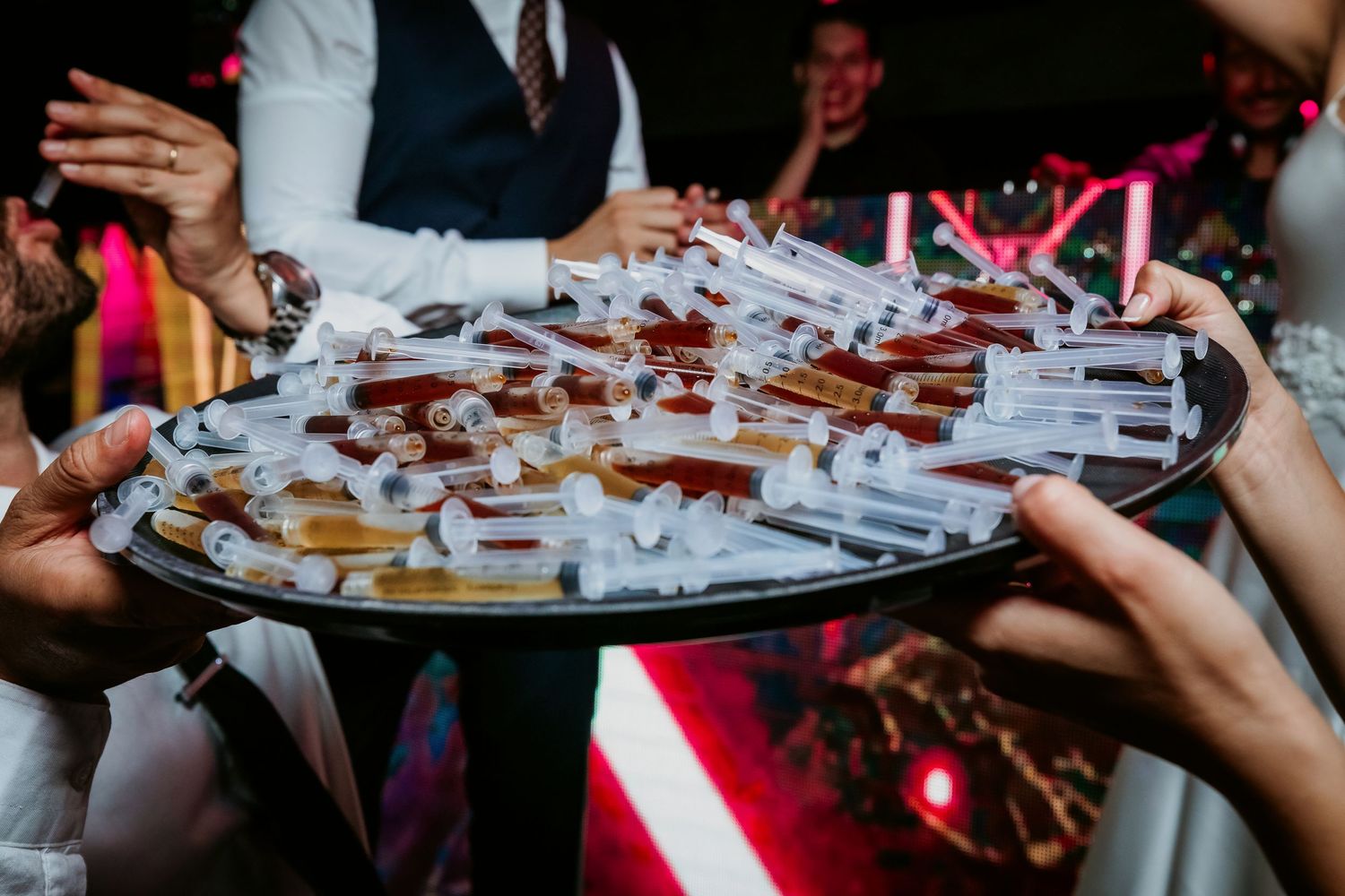 Guests reach for syringes filled with colorful drinks on a tray at a wedding party.