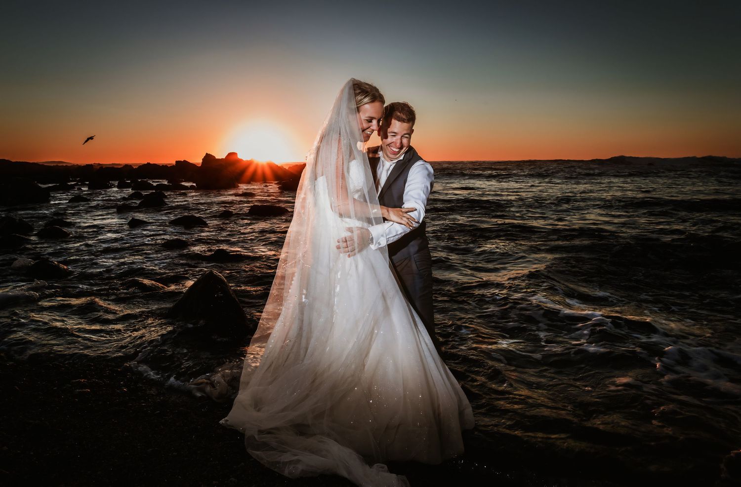 Newlywed couple embracing on rocky shoreline with a stunning sunset over the ocean behind them.