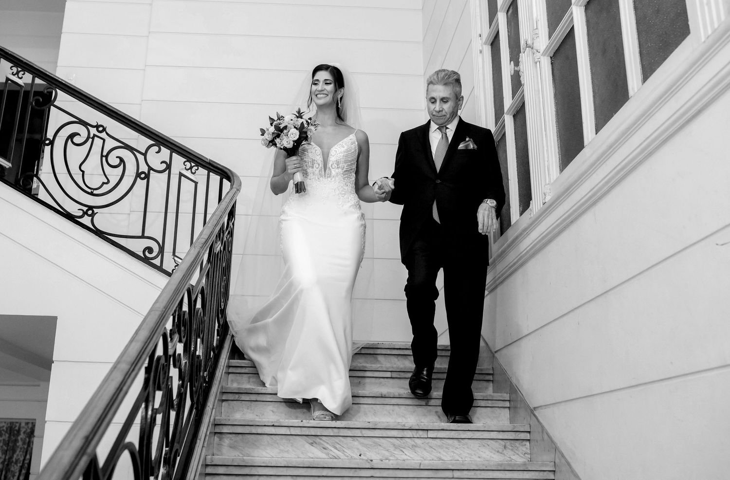 Black and white photo of a bride in a white gown descending stairs with an older man in a suit.
