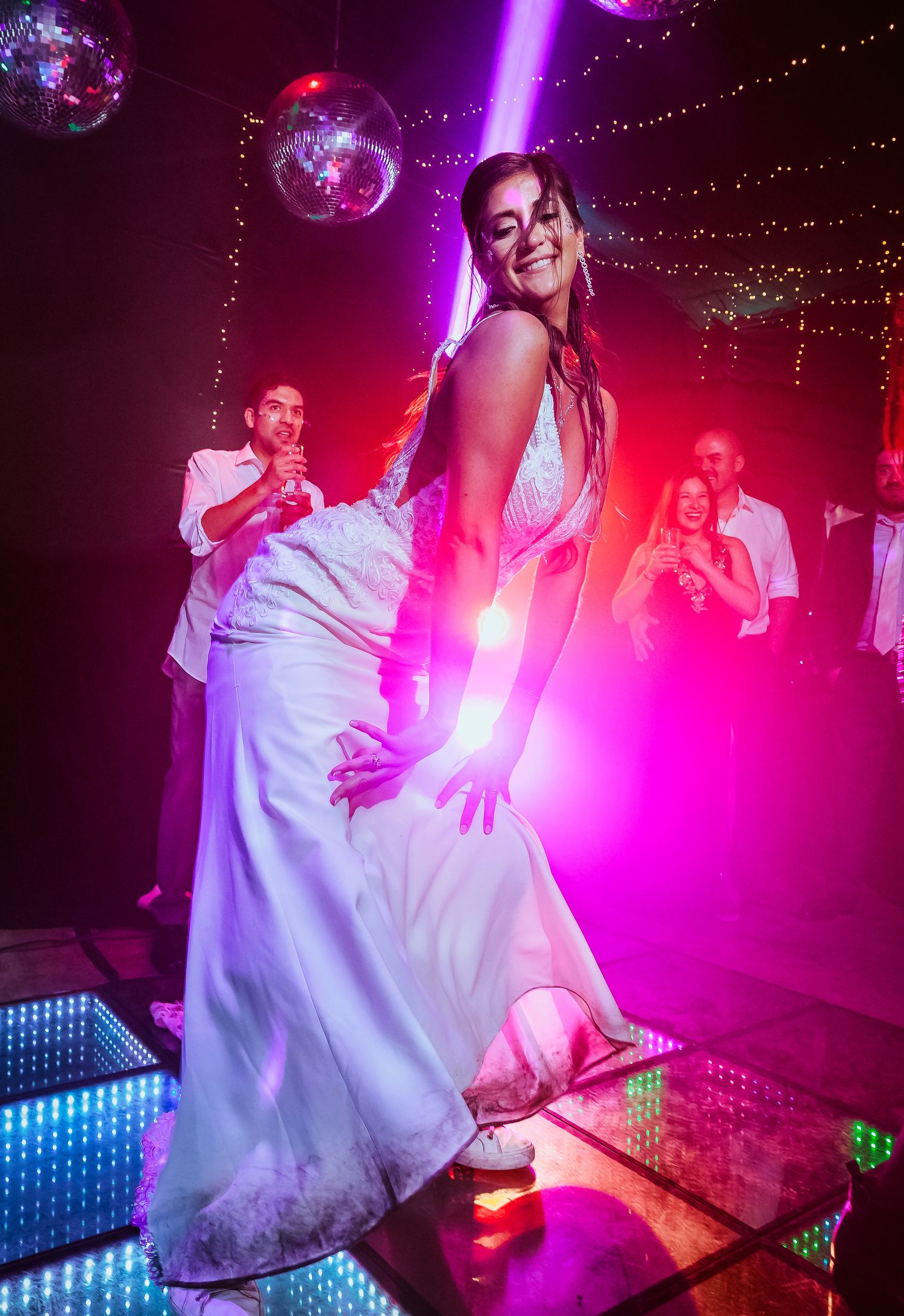Bride in white gown dancing joyfully on illuminated dance floor with disco balls and colorful lights at wedding reception.