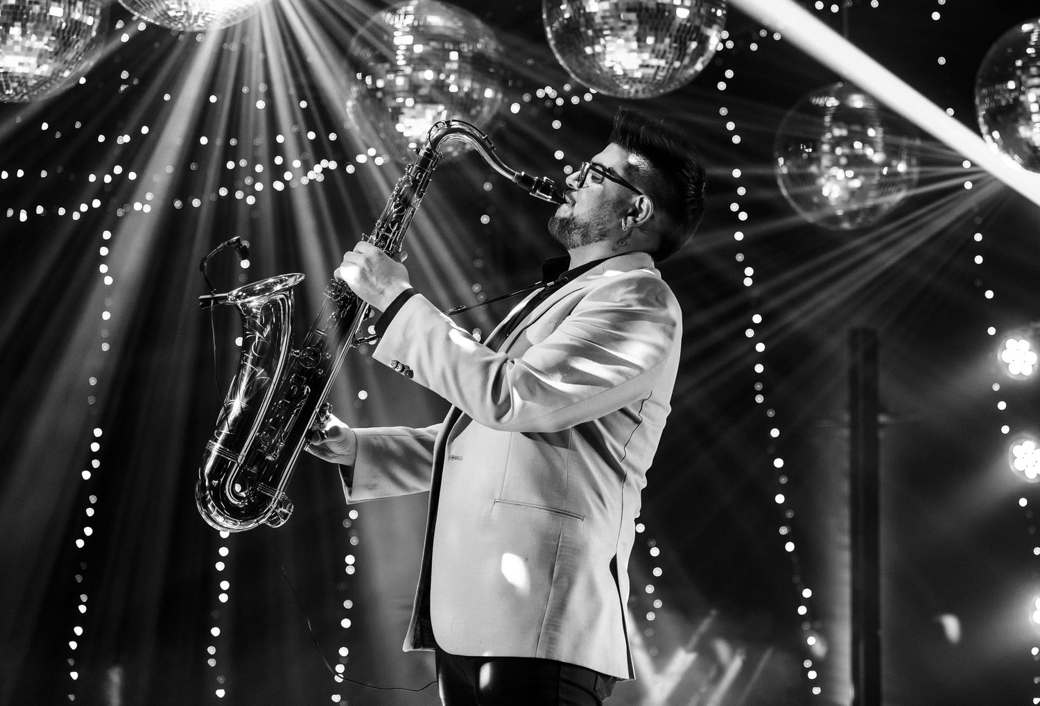 Man in white tuxedo playing saxophone on stage with dramatic disco ball lighting in black and white photo.
