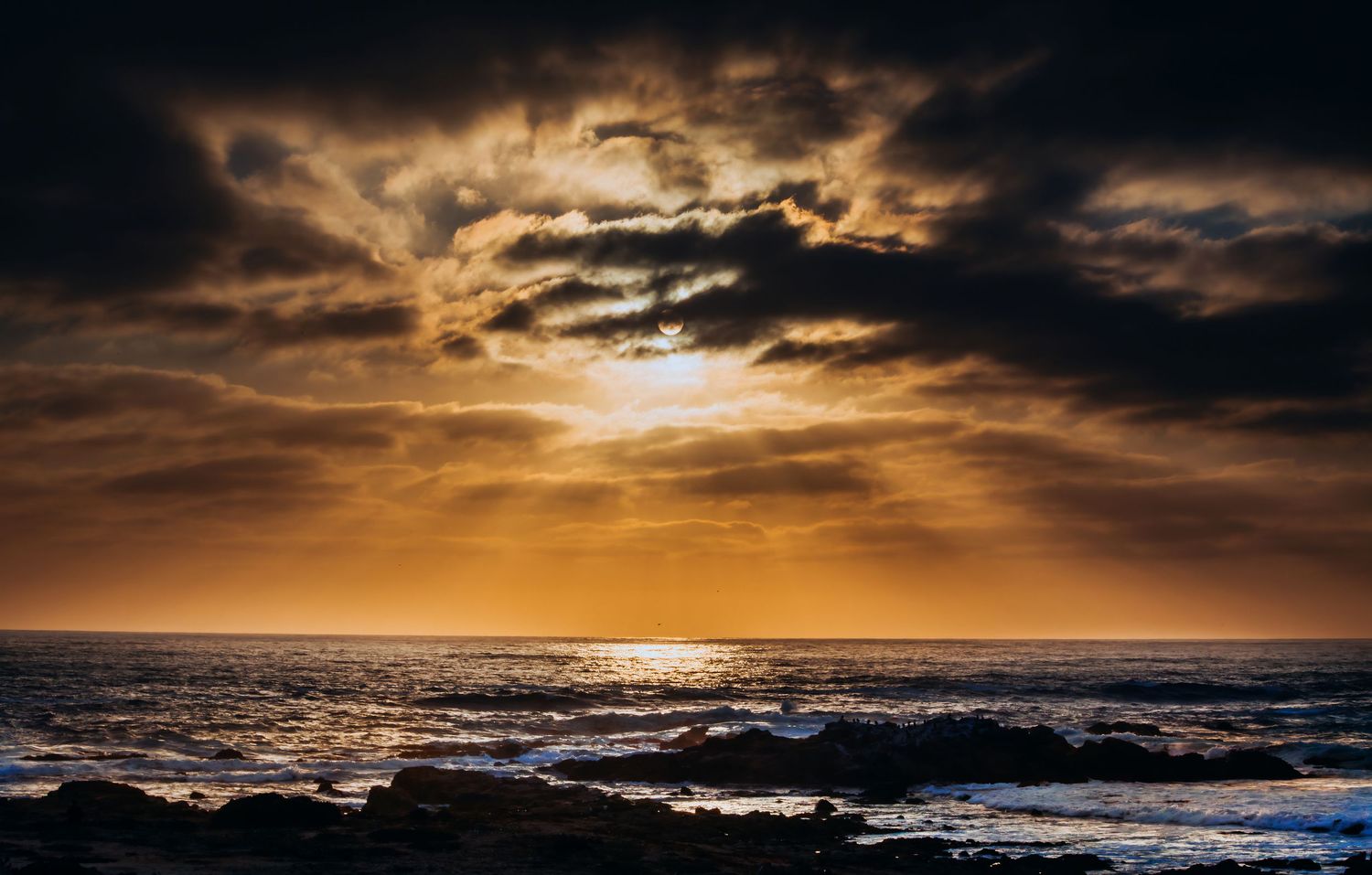 Dramatic ocean sunset with golden rays breaking through dark storm clouds over rocky shoreline.