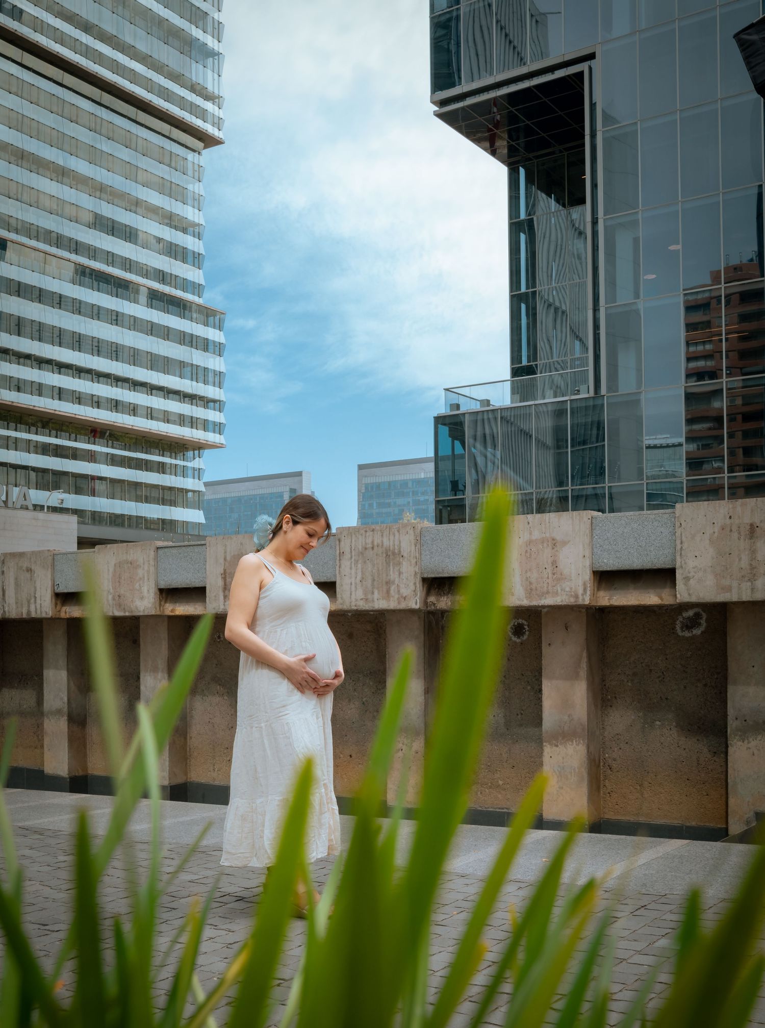Pregnant woman in a white dress holding her belly while standing between tall modern city buildings under blue sky.