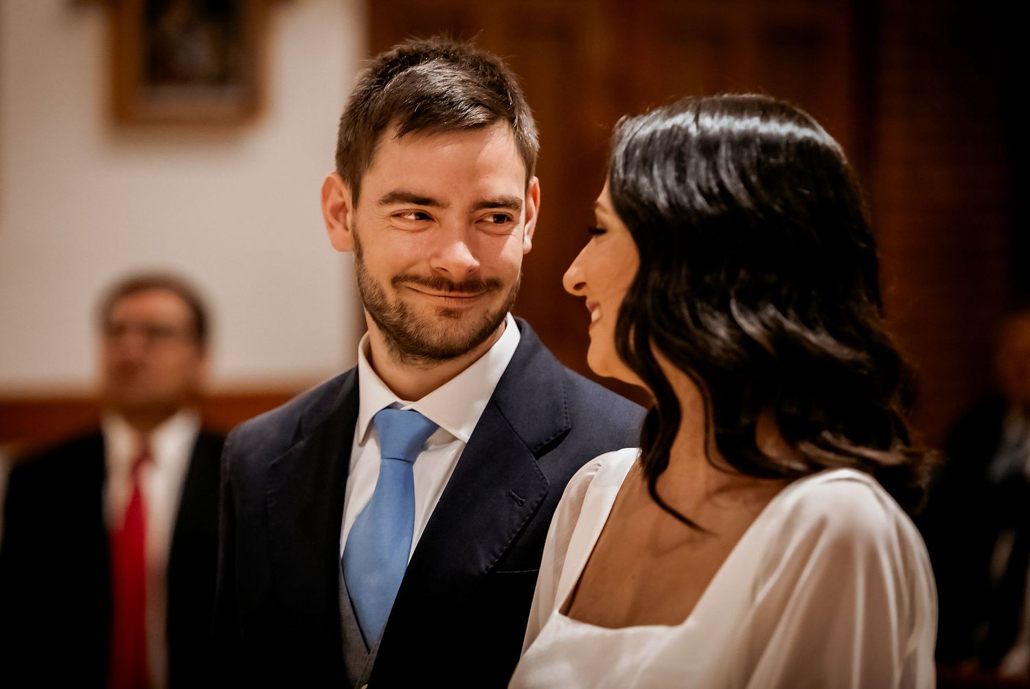 Groom in navy suit with blue tie smiling at bride in white dress during wedding ceremony.