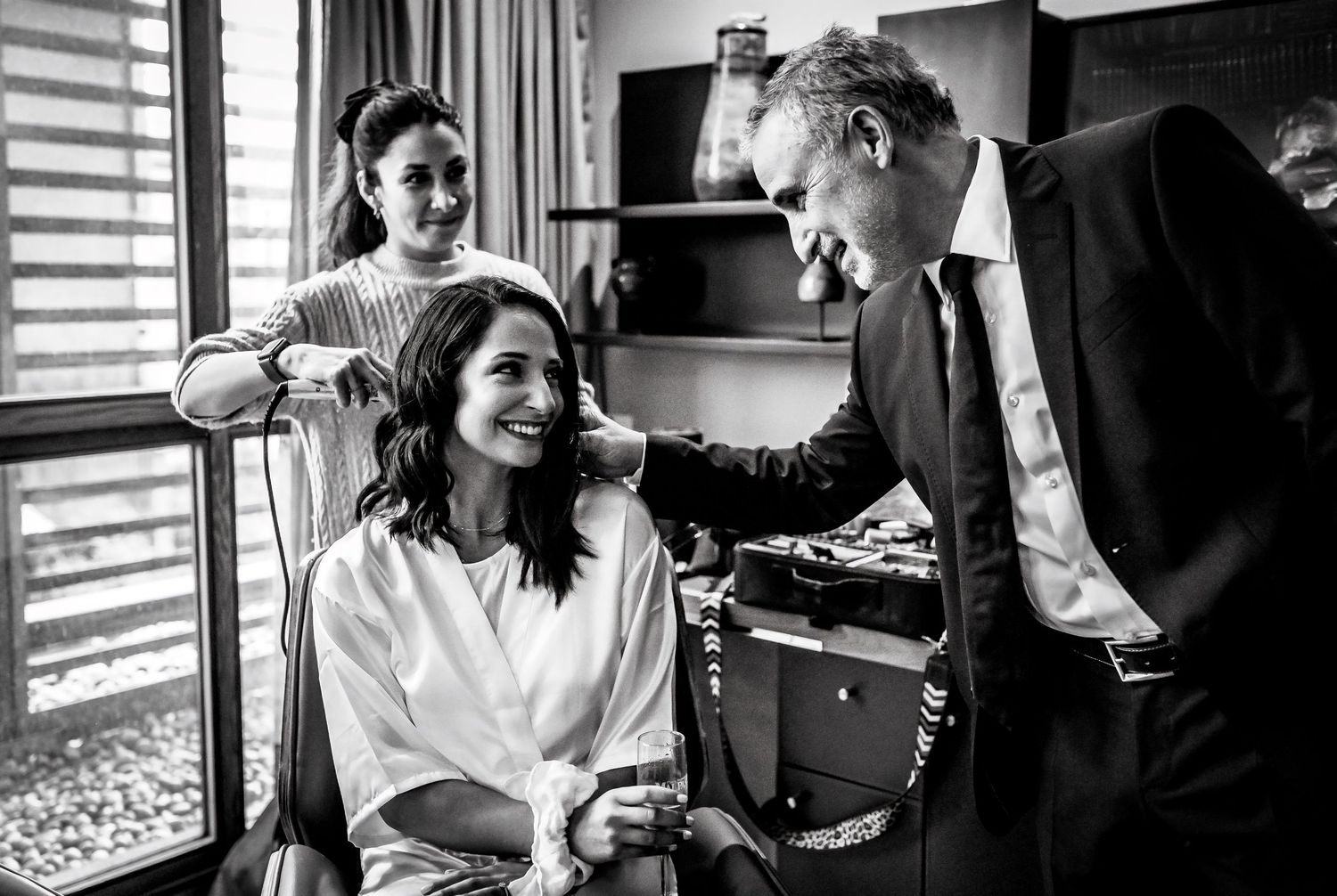 Black and white photo of a bride getting hair done while a man in a suit smiles at her during wedding preparations.