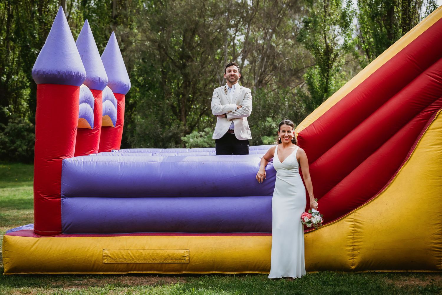 Bride and groom posing in front of a colorful bouncy castle at their outdoor wedding reception.