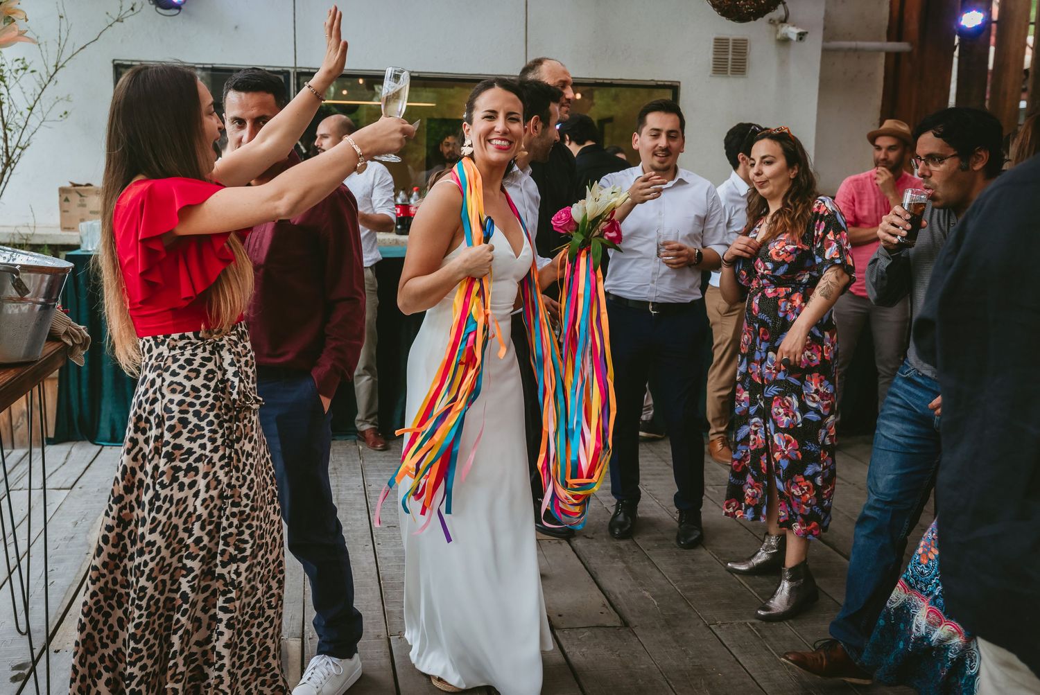 Bride in white dress with colorful ribbons dancing joyfully with guests at a vibrant wedding reception party.