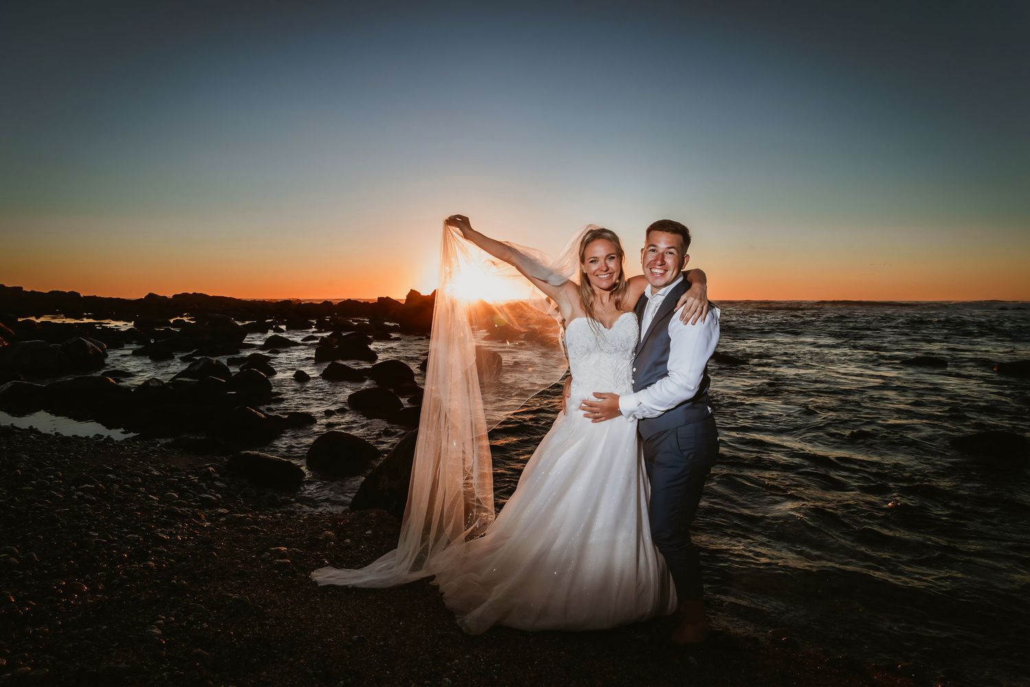 Joyful couple on rocky beach at sunset, bride holding her flowing veil up high.