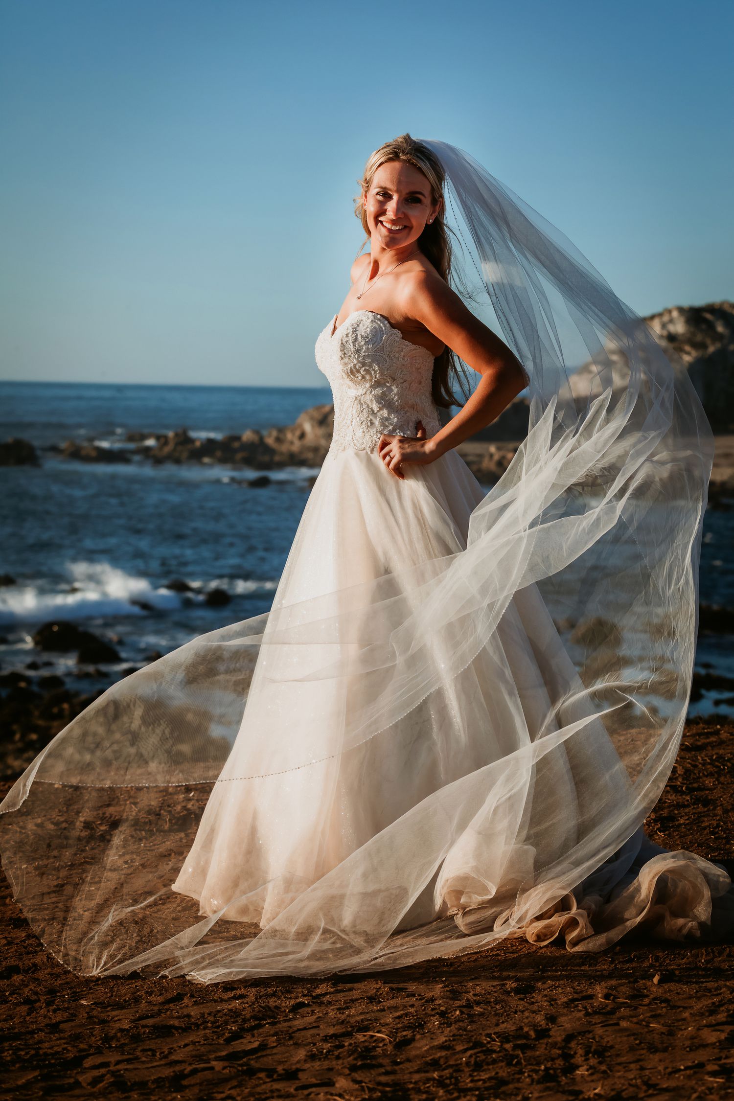 Radiant bride in strapless wedding dress with dramatic veil blowing in wind on rocky ocean beach.
