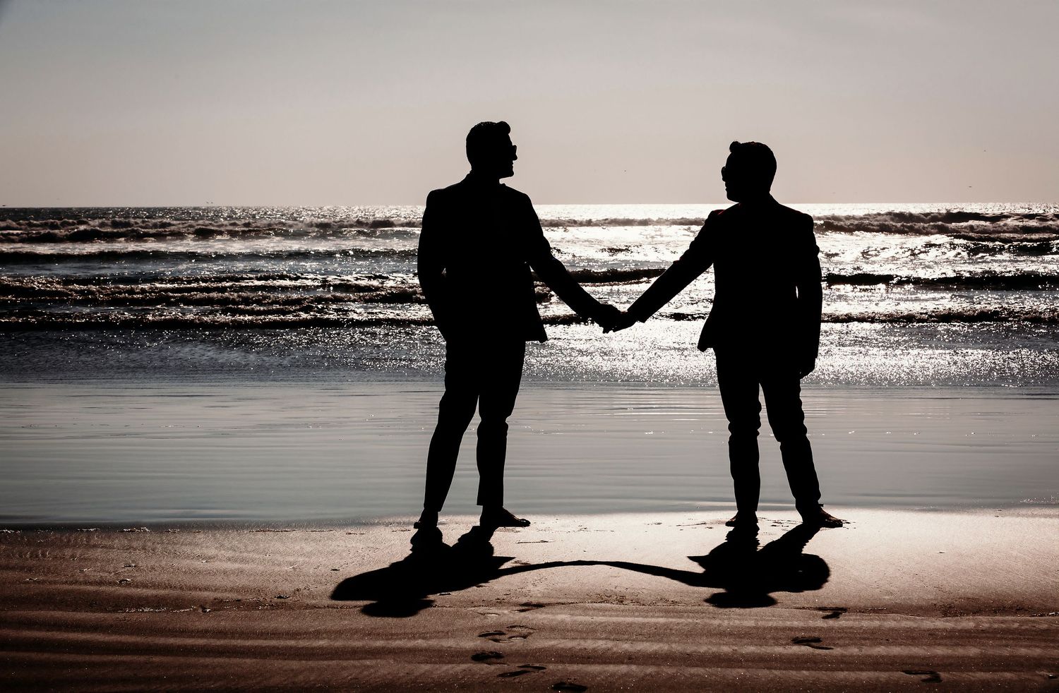 Romantic silhouette of two grooms holding hands on a beach at sunset with ocean waves behind them.