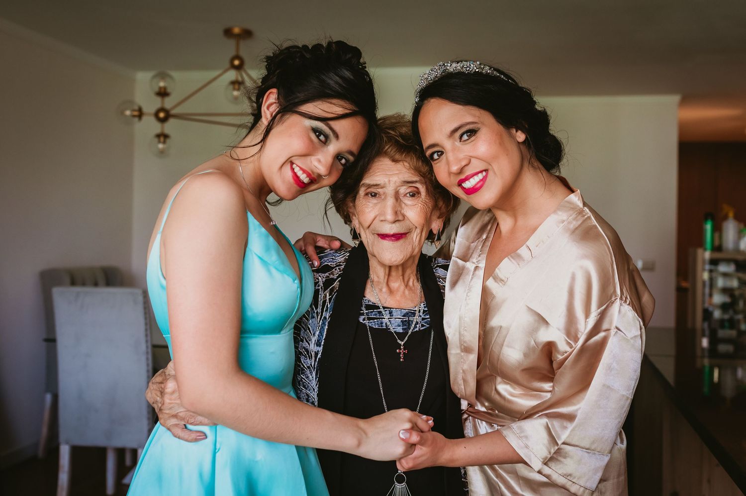 Two smiling women in formal attire embracing elderly grandmother before wedding celebration.