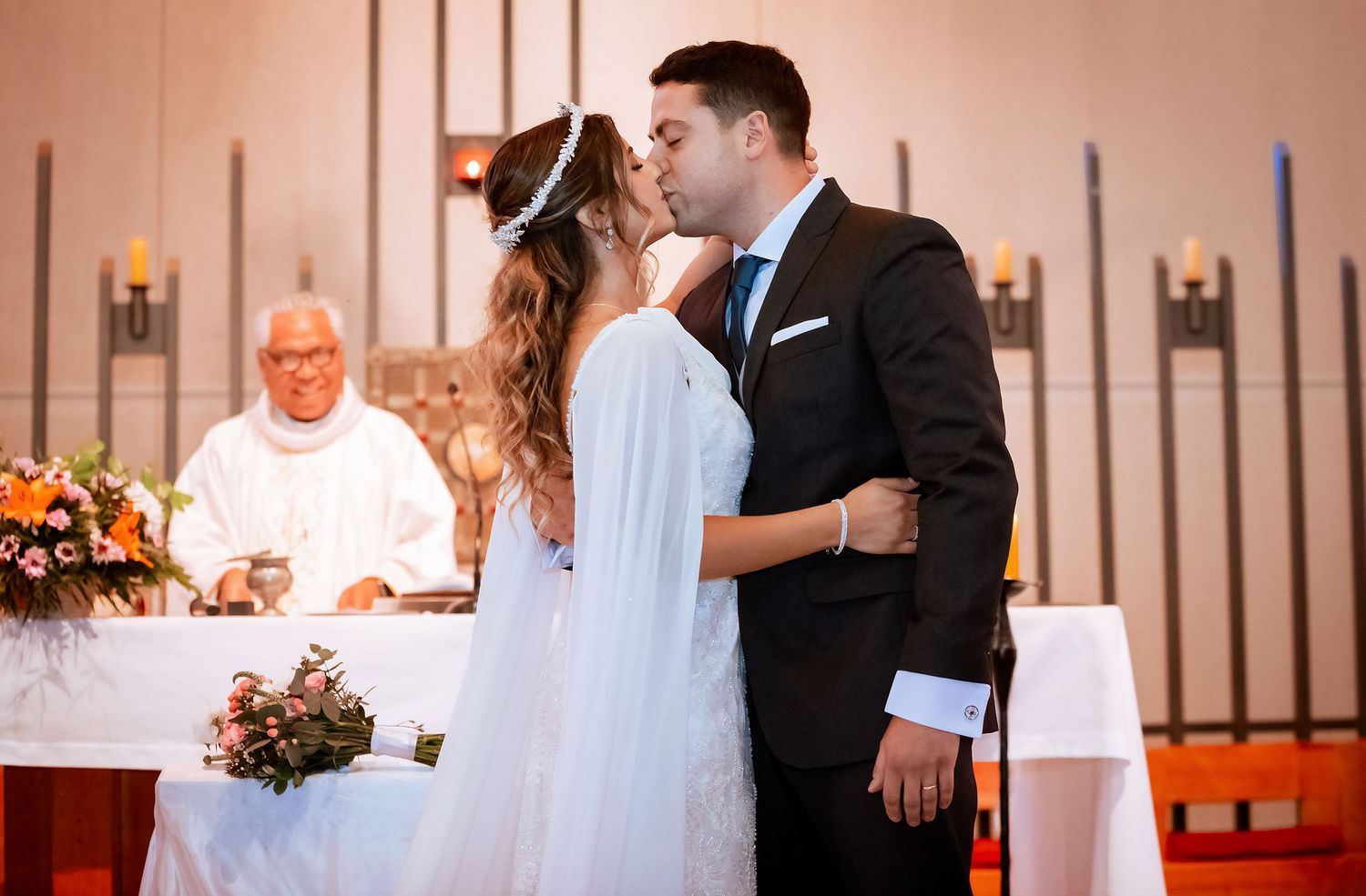 Bride and groom sharing first kiss at church altar while priest smiles in background during wedding.
