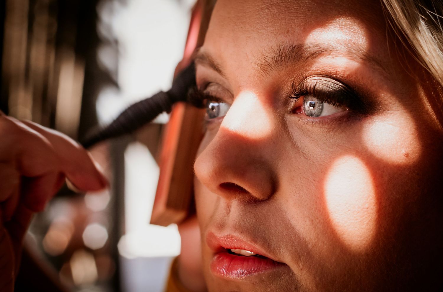 Close-up of bride getting eye makeup applied by makeup artist with soft dappled sunlight on her face.