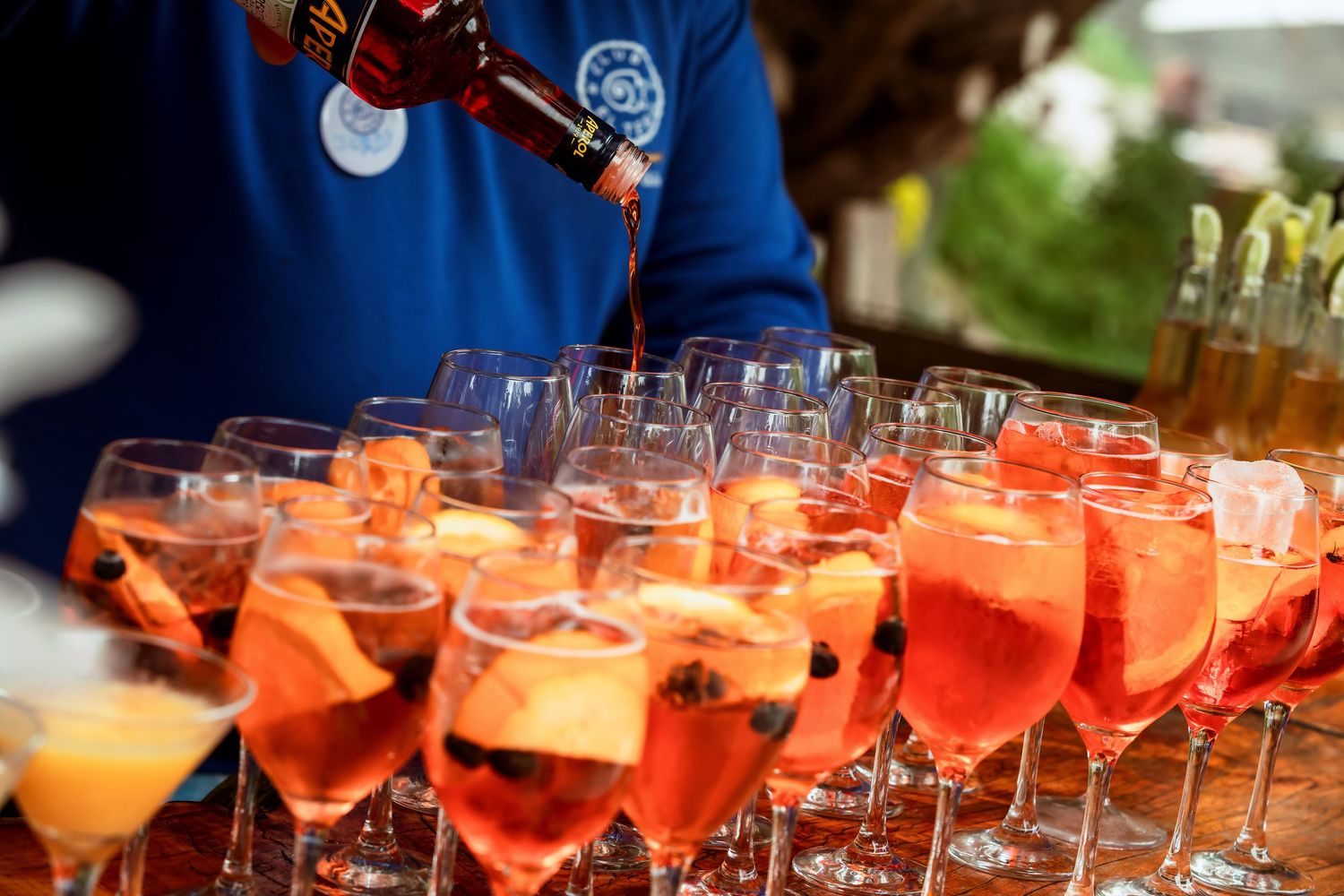 Bartender pours red aperol spritz cocktails into elegant wine glasses at an outdoor wedding reception.