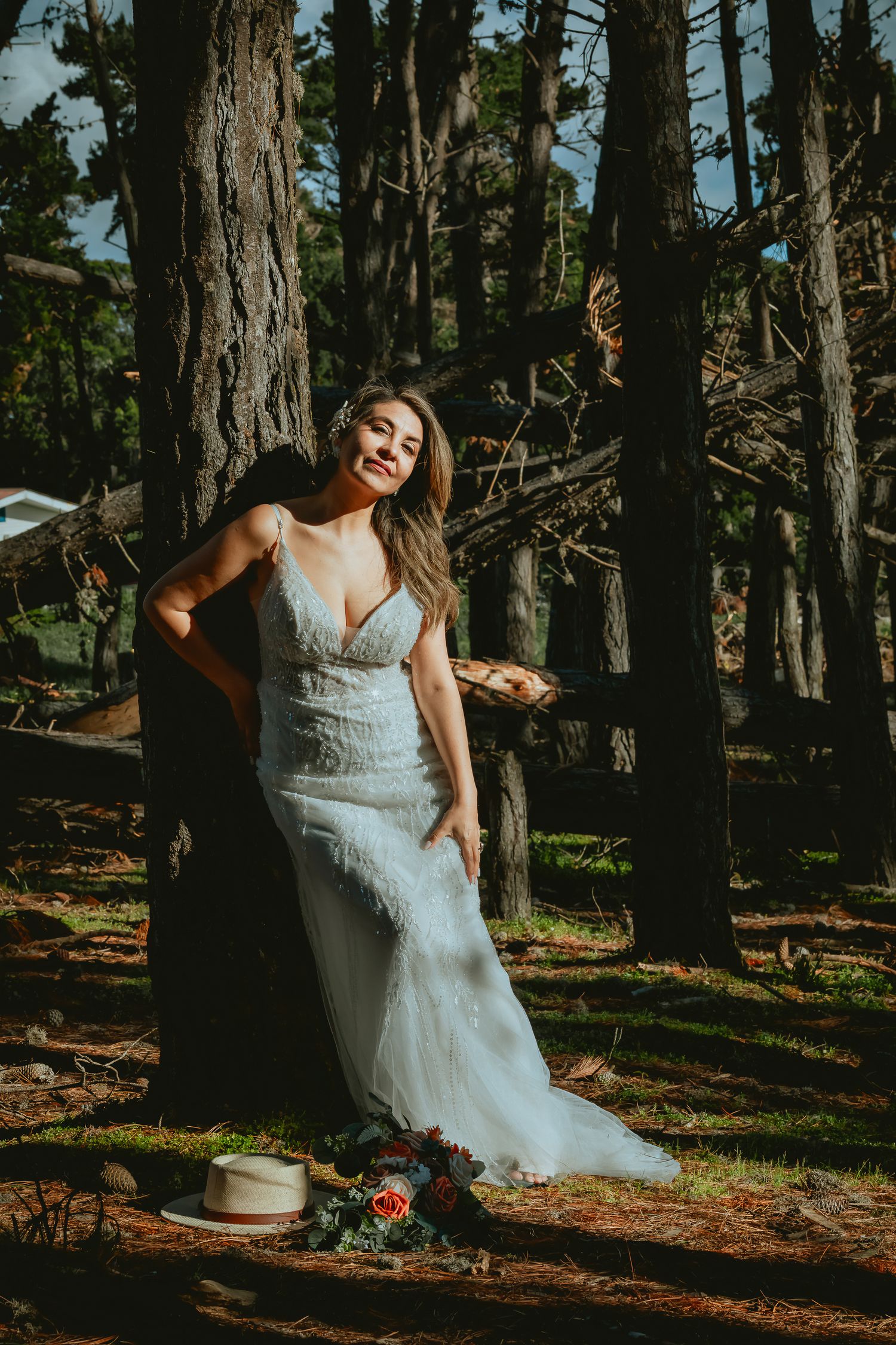 Bride in a lace wedding gown leans against a tall pine tree in a sunlit forest, with a straw hat and bouquet resting on the ground.